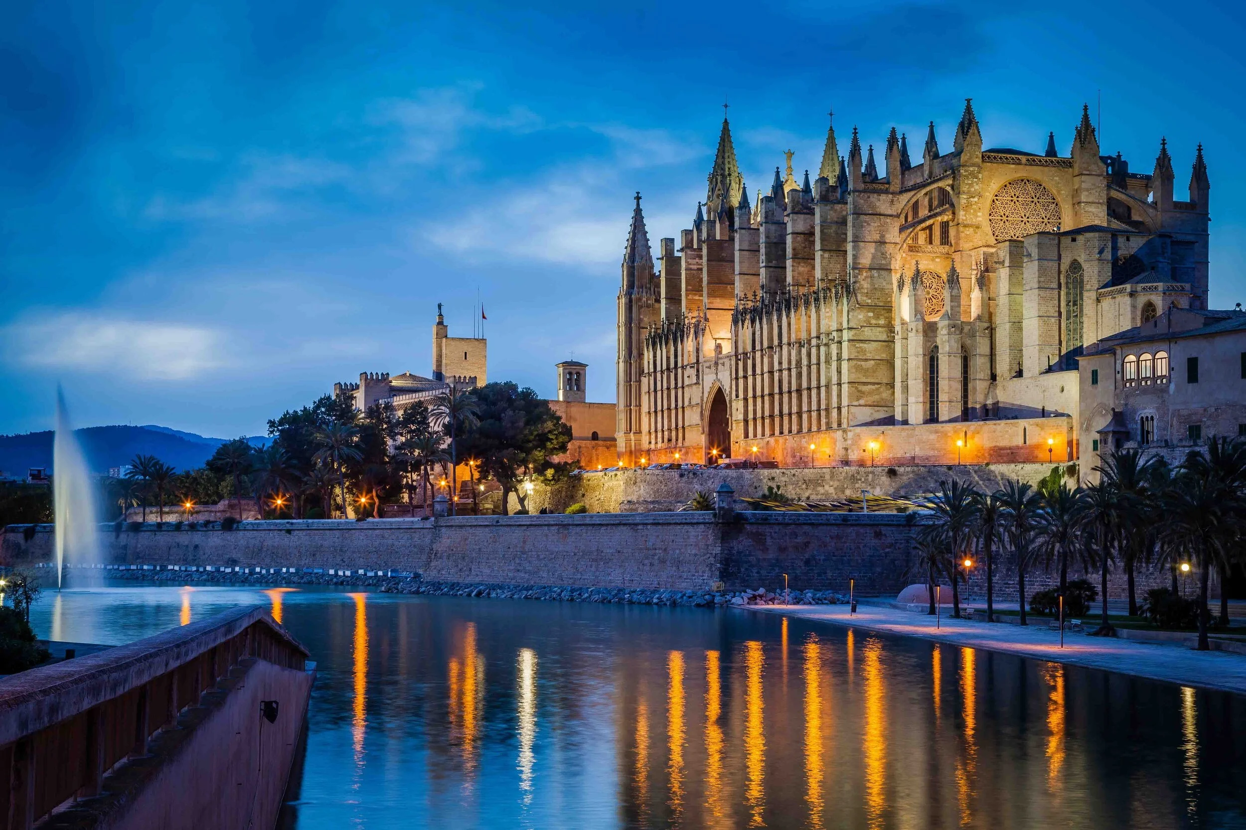 Catedral de Palma de Mallorca iluminada por la noche, vista desde el agua con reflejos en el río y un fountain en primer plano.