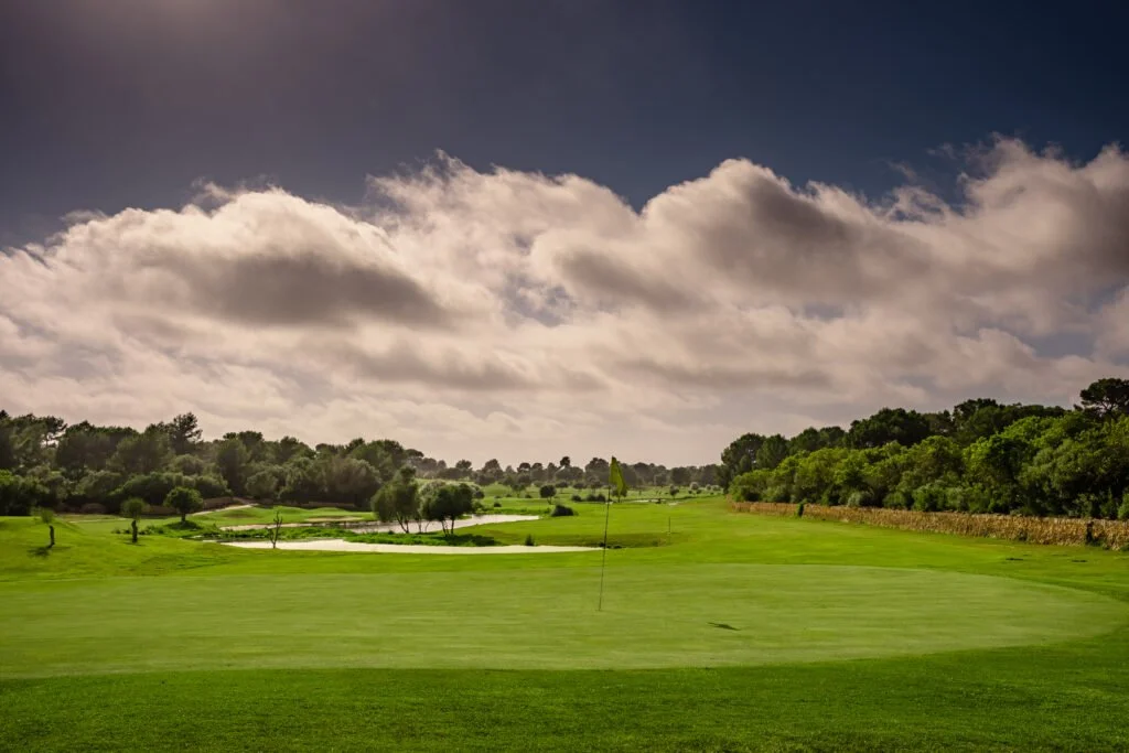 Campo de golf con césped verde, obstáculos de agua, árboles y un cielo con nubes.