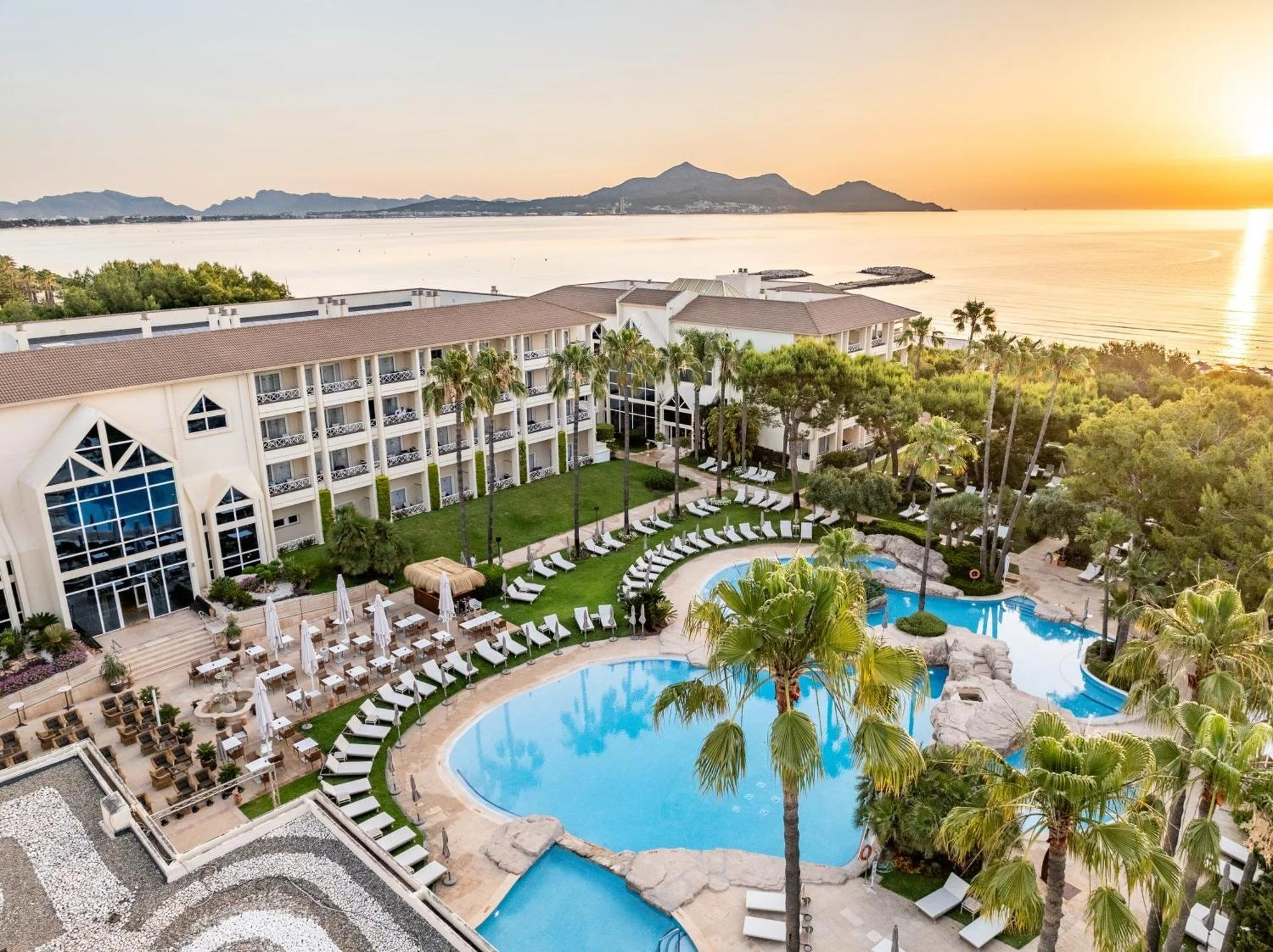 Vista de un complejo hotelero con piscina, palmeras y habitaciones, con un cuerpo de agua y montañas al fondo durante el atardecer.