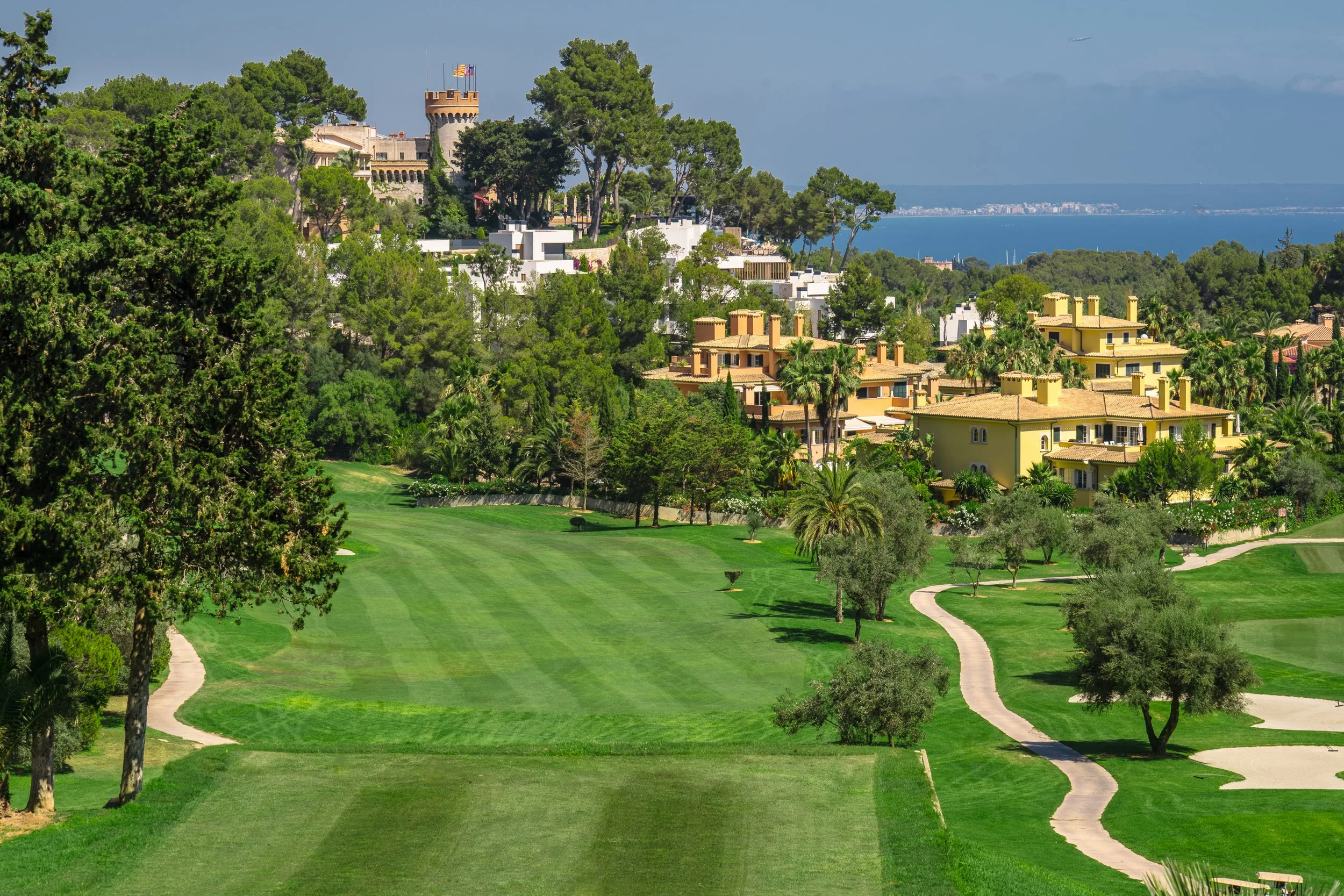 Campo de golf con casas residenciales y árboles verdes, vista hacia el mar y un castillo en la colina, cielo despejado.