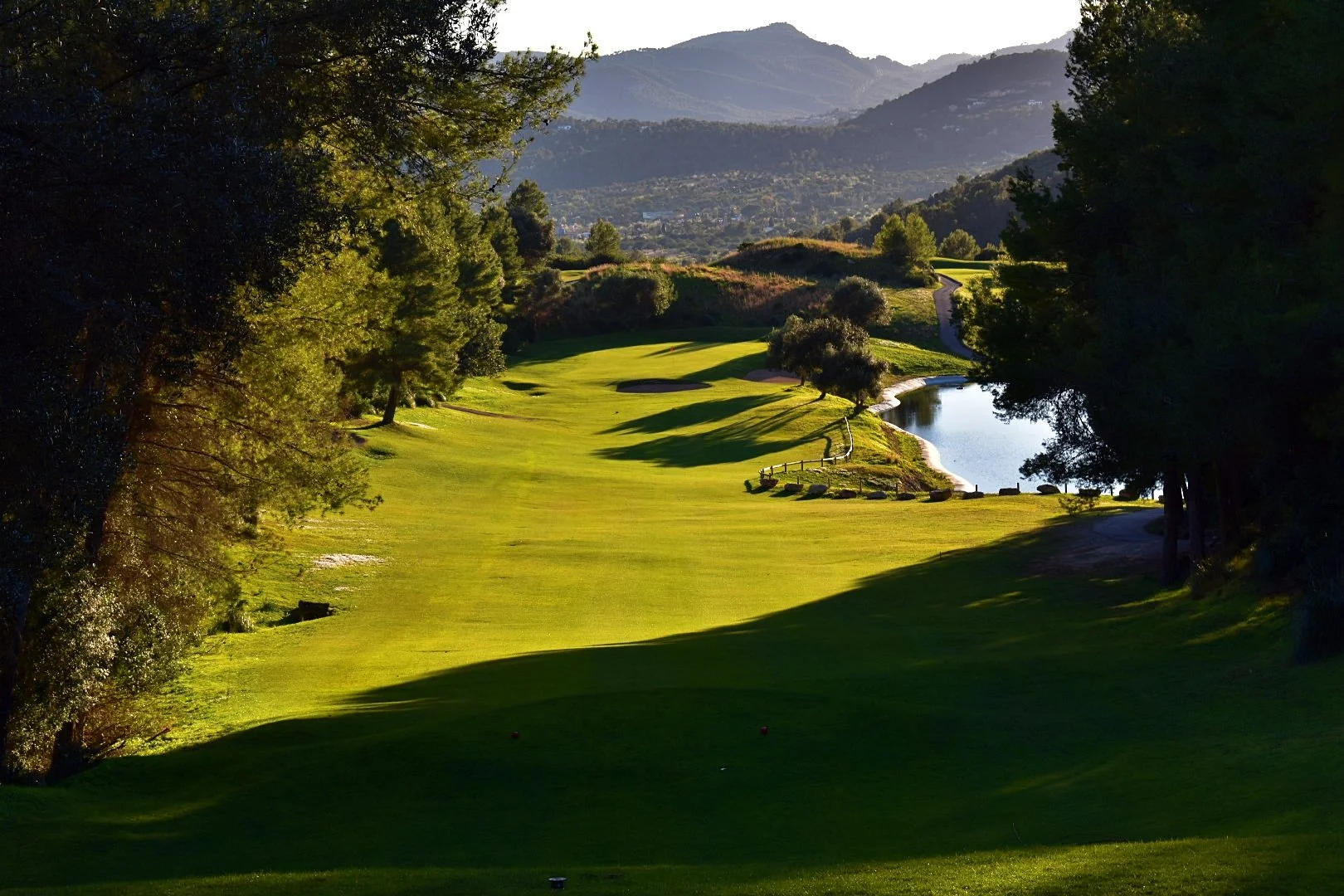 Campos de golf con árboles y un pequeño lago, montañas al fondo.