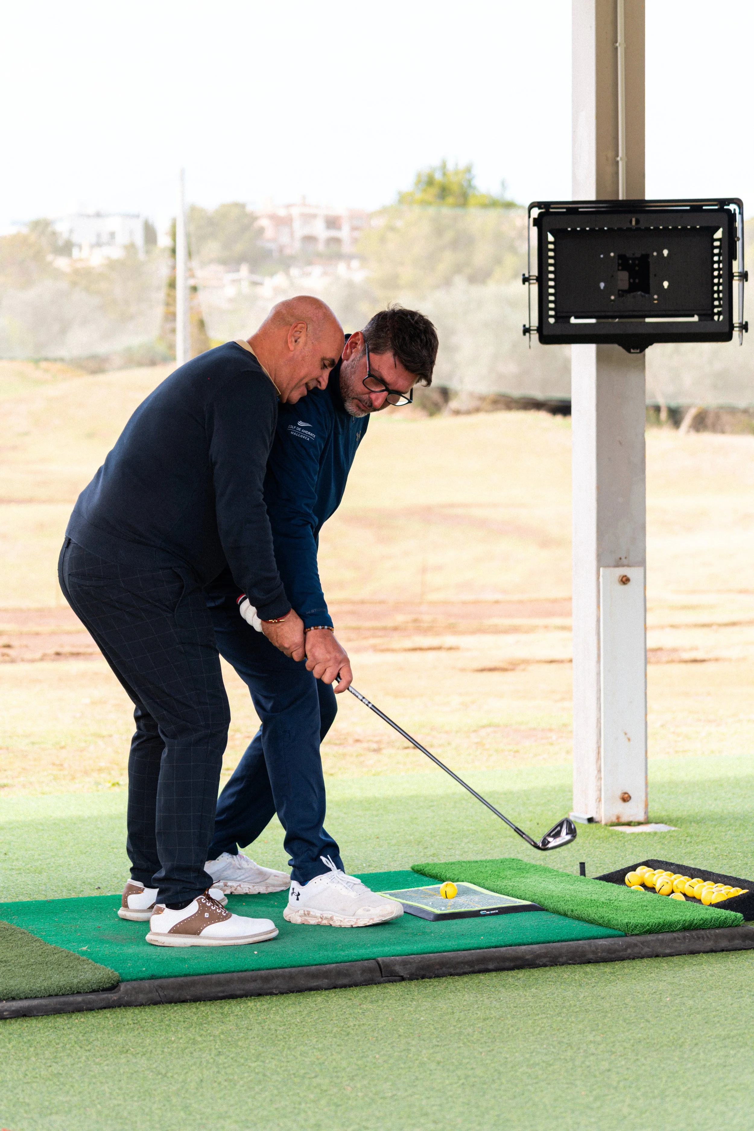 Dos hombres practicando golf en un campo de golf cubierto, uno ayudando al otro a colocar la bola en el tee.