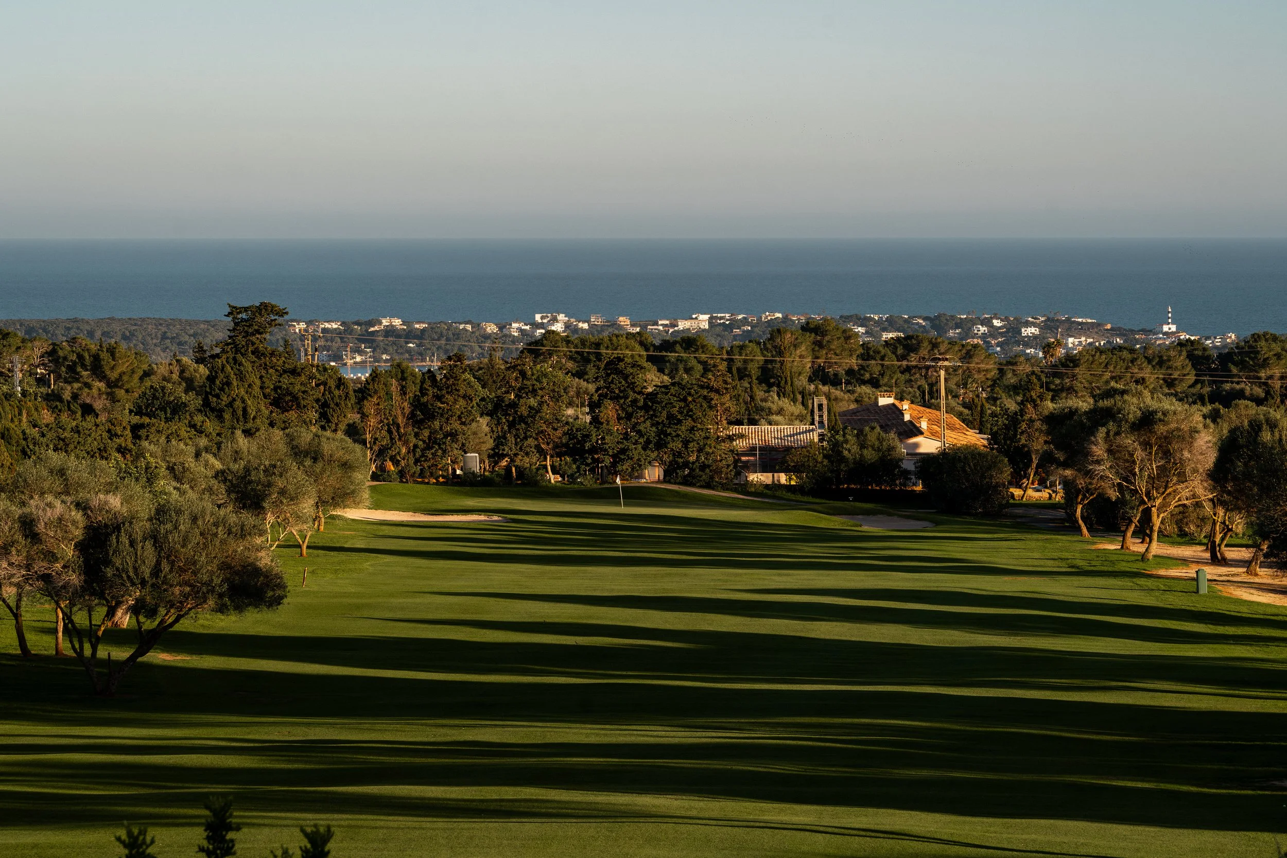 Campo de golf con árboles a los lados, casas en el fondo y el océano en el horizonte.