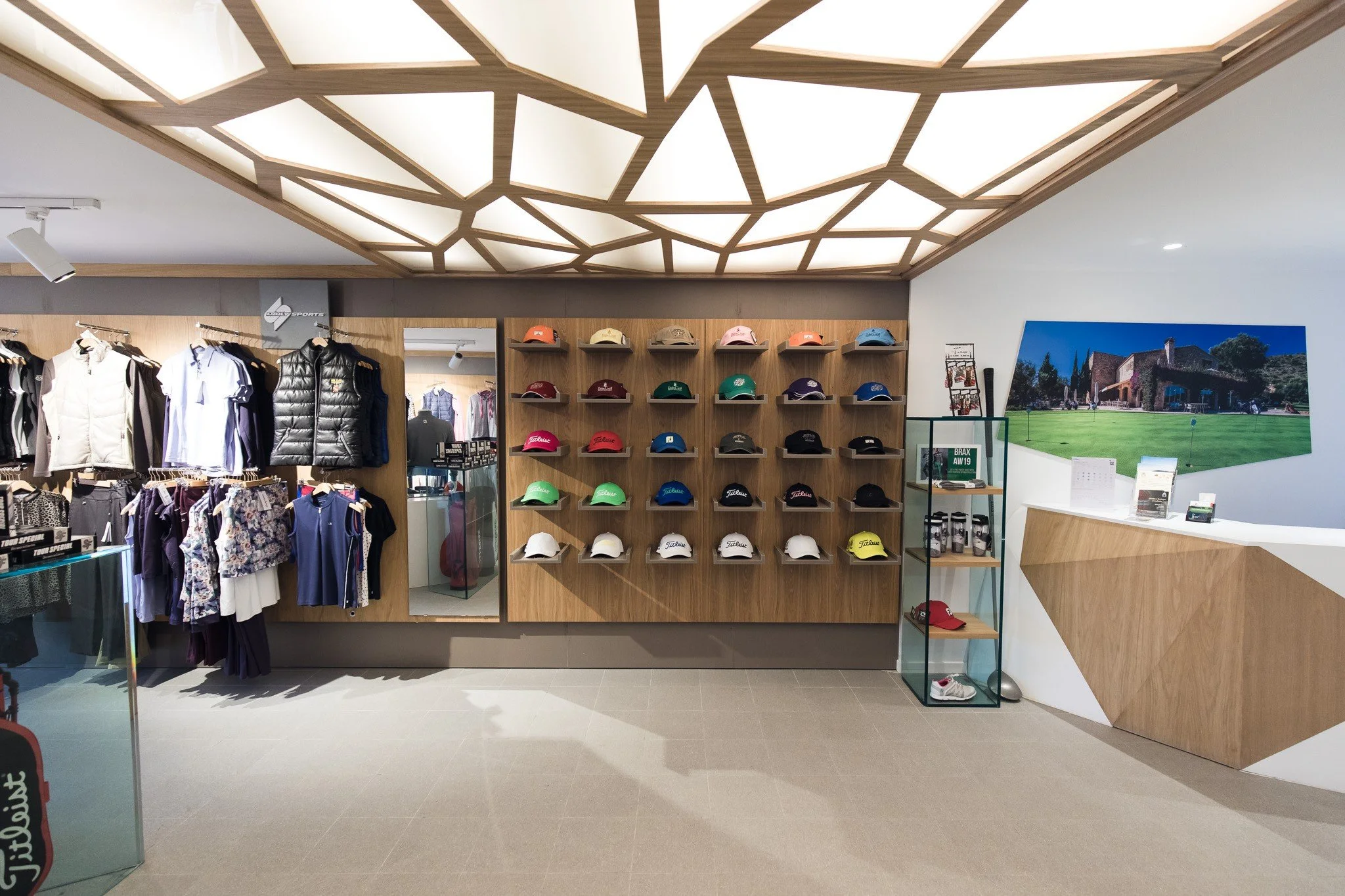 Interior de una tienda de artículos deportivos, con gorras de diferentes colores en una pared de madera, ropa en exhibición y una imagen grande de un campo de golf.