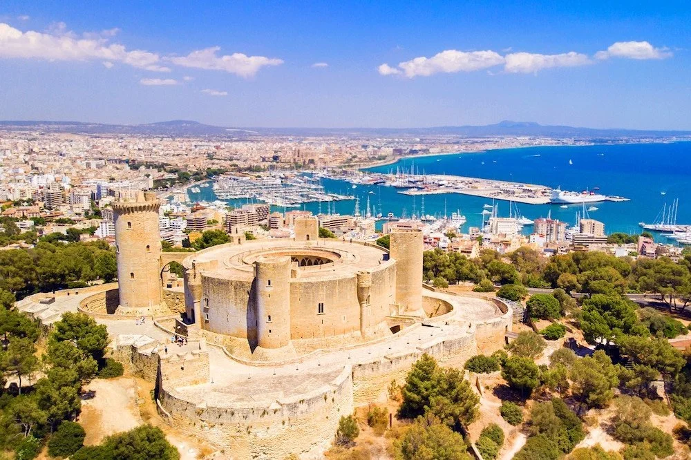 Vista aérea de un castillo medieval en el centro de una ciudad junto al mar, con un puerto lleno de yates y barcos.