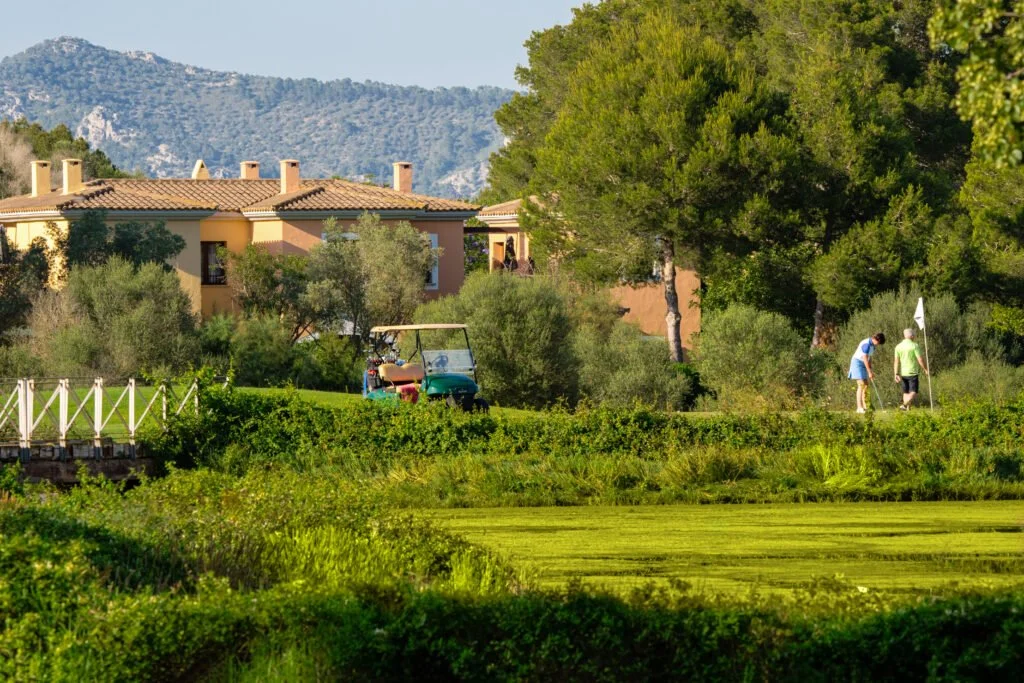 Personas jugando golf en un campo con casas y montañas de fondo.