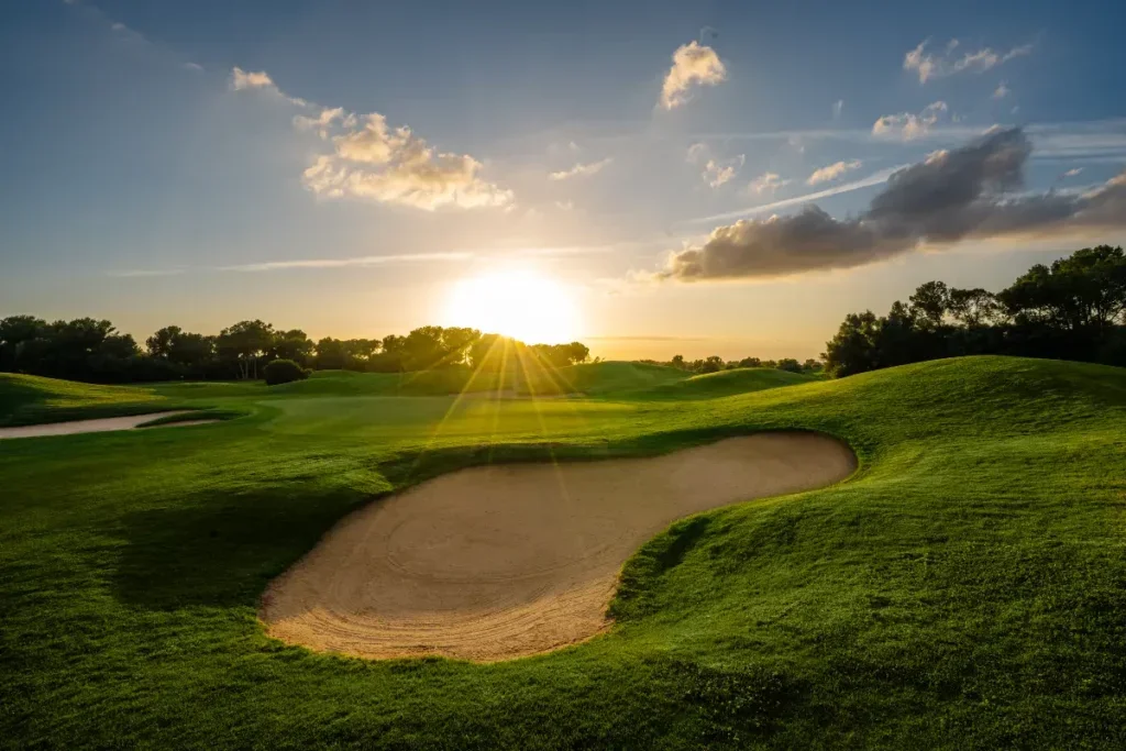 Campo de golf con búnker y césped verde bajo un cielo con nubes y sol en el horizonte