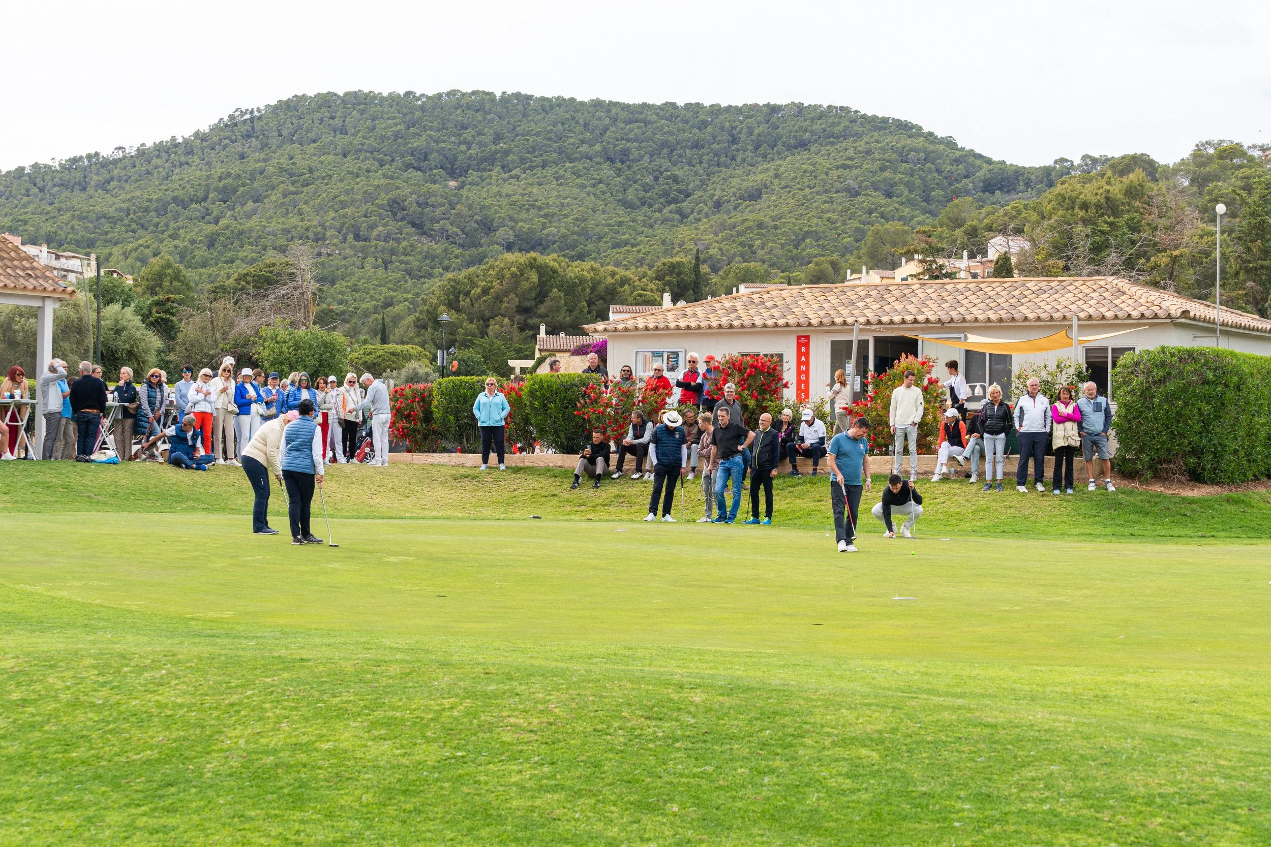 Grupo de personas jugando golf en un campo verde, con casas y colinas verdes al fondo y árboles alrededor.