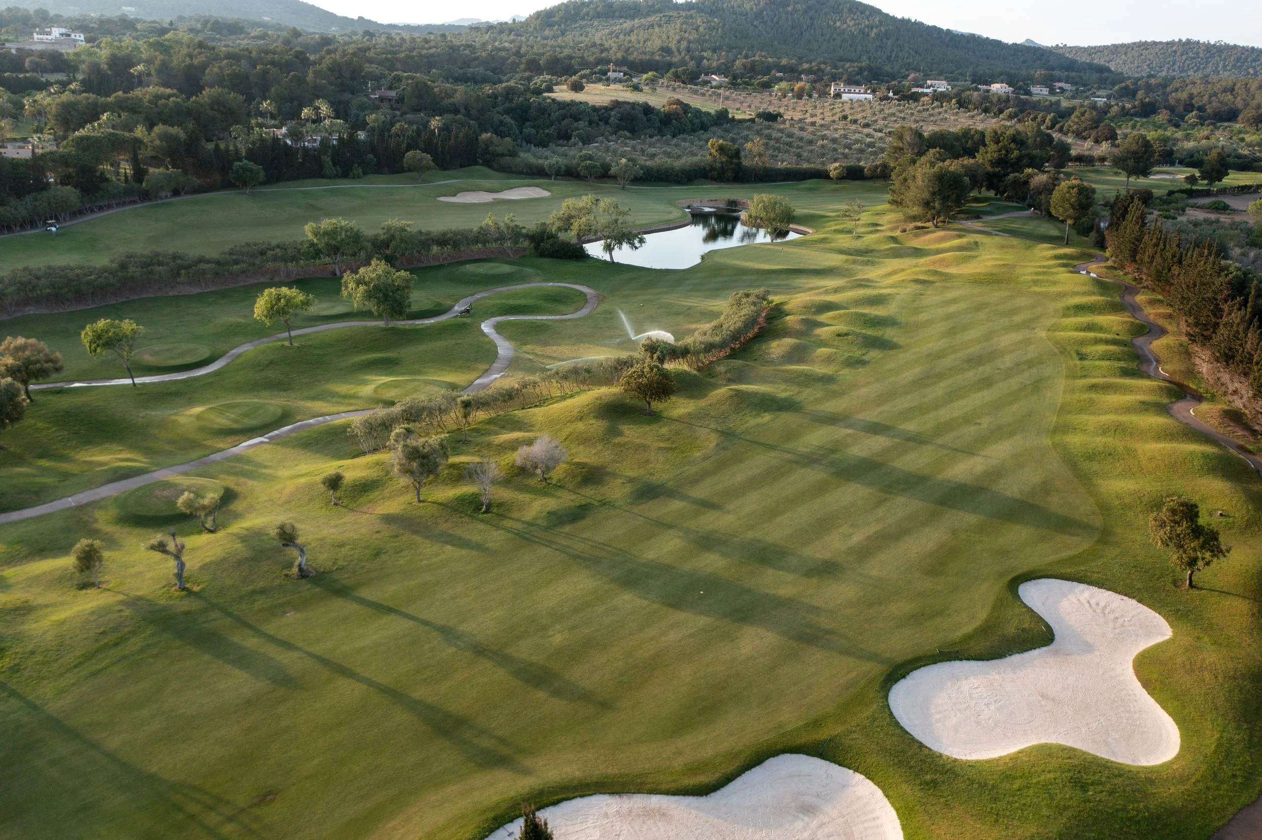 Vista aérea de un campo de golf con áreas verdes, búnkeres de arena, árboles dispersos y un pequeño cuerpo de agua, rodeado de colinas y vegetación.