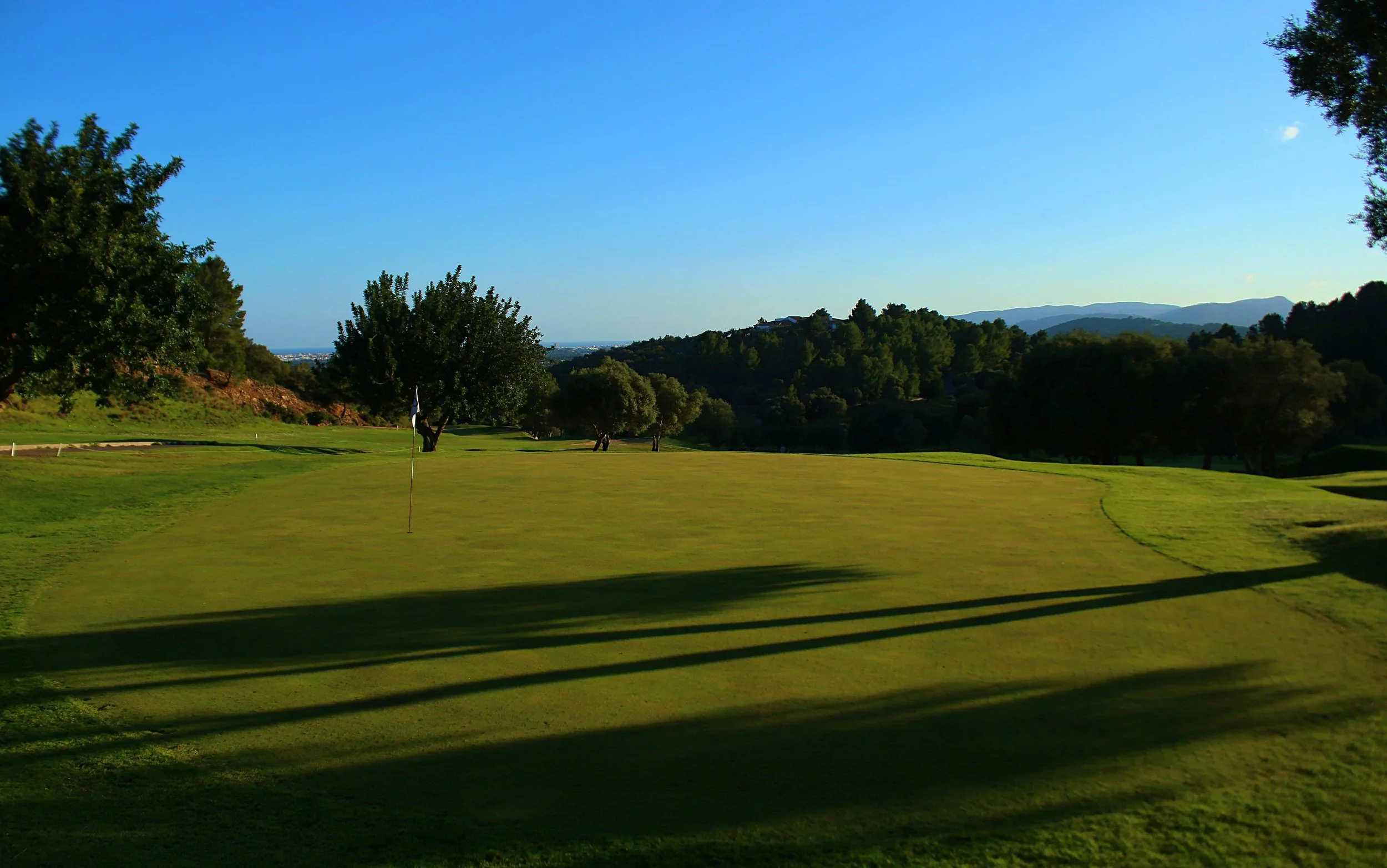 Vista de campo de golf con árboles y colinas en el fondo, bajo un cielo despejado.