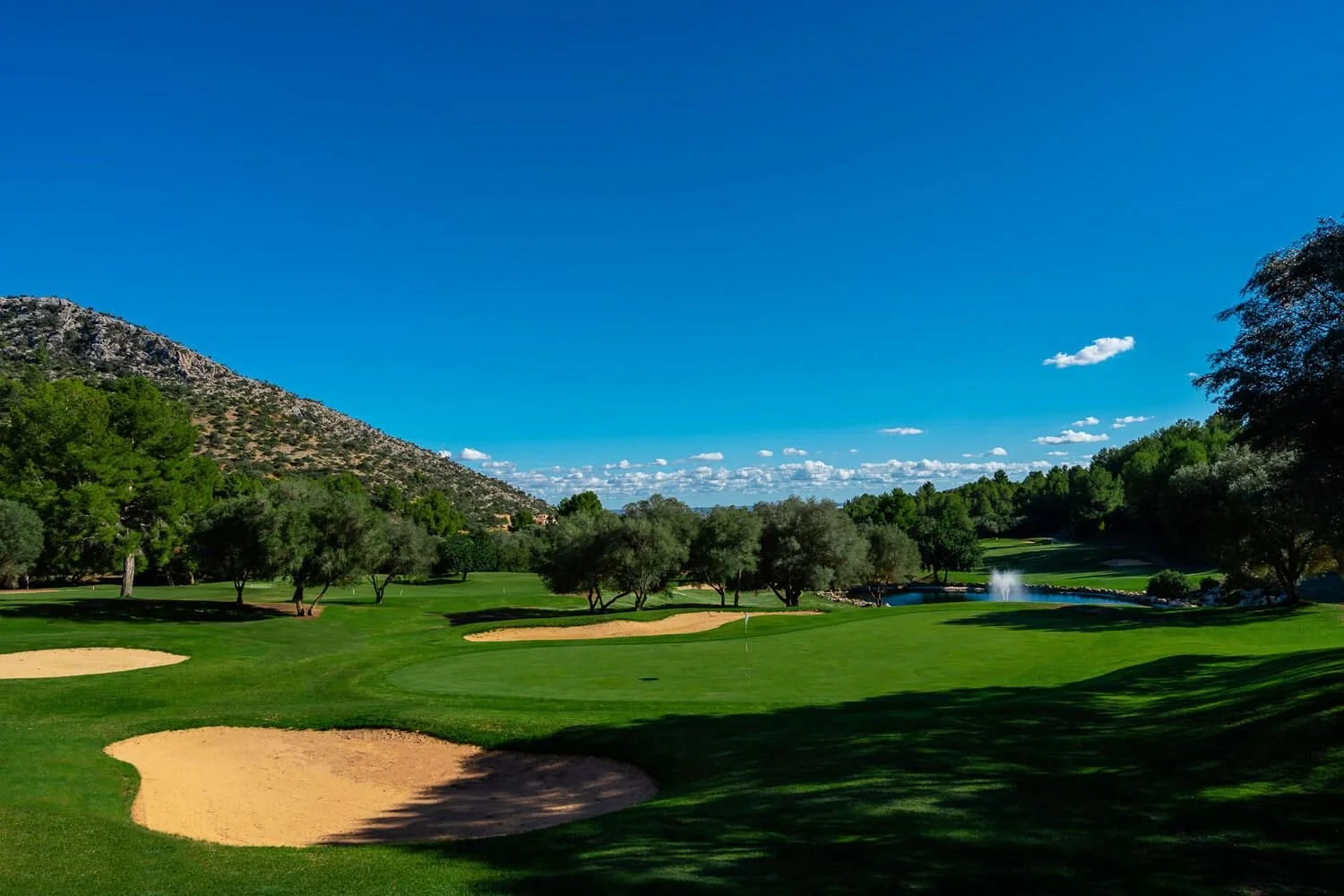 Campo de golf con árboles, arena y un estanque bajo un cielo azul con nubes en un paisaje montañoso.
