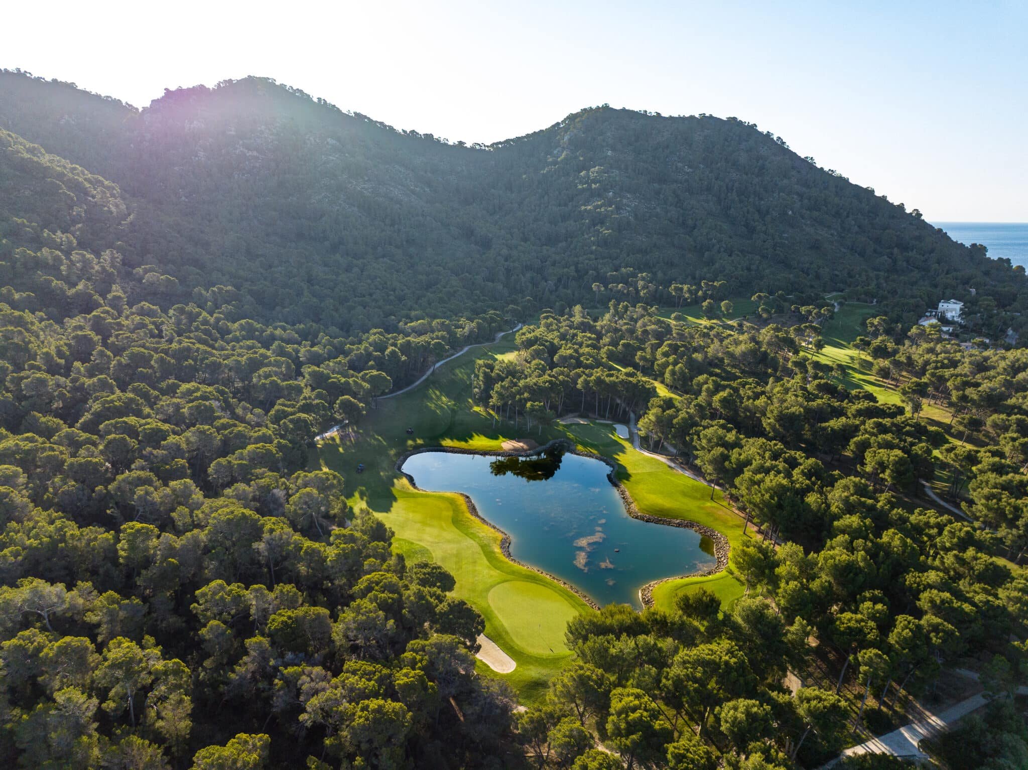 Vista aérea de un campo de golf con un estanque en medio rodeado de árboles y montañas al fondo, con el sol iluminando la escena.