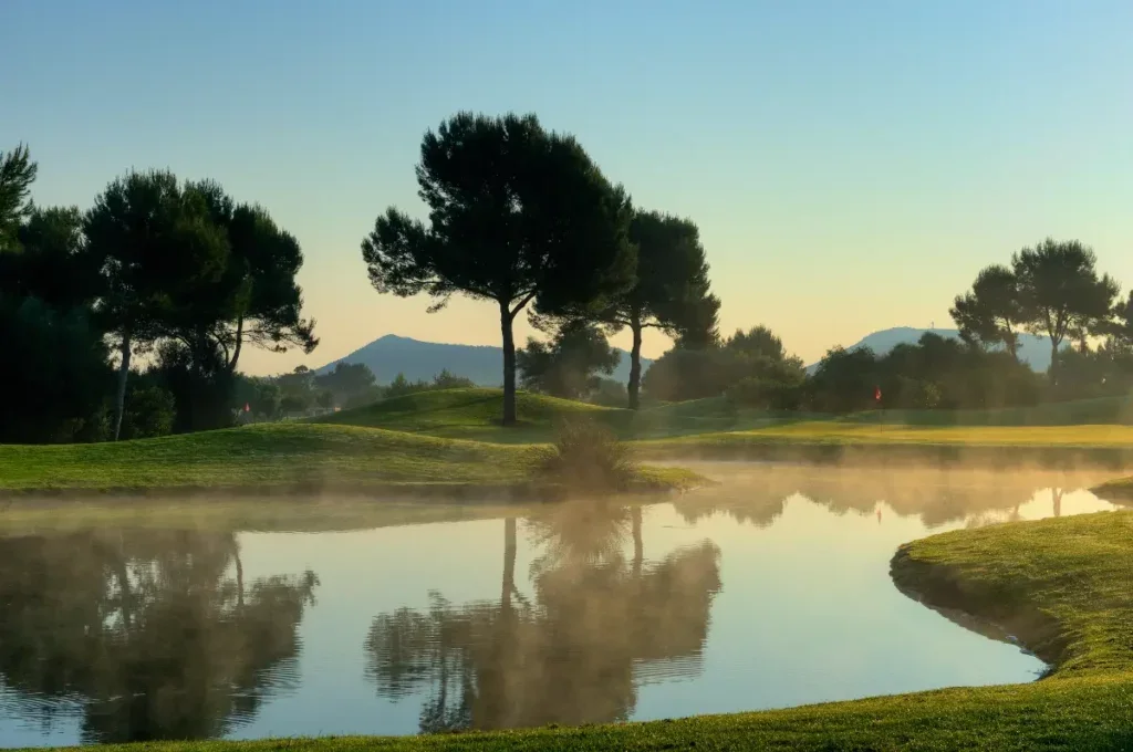 Paisaje de un campo de golf con árboles y un lago, con neblina matutina y Montañas al fondo.