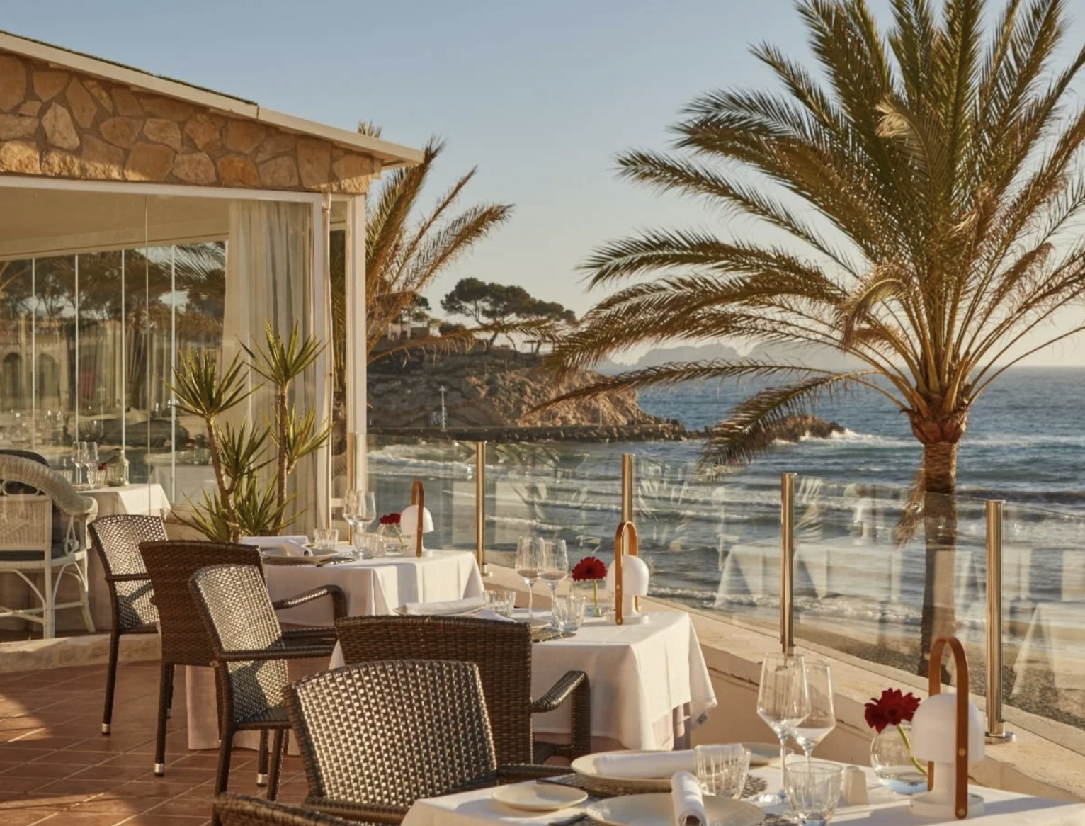 Mesas de restaurante con manteles blancos, copas y flores rojas, frente a una vista del mar con palmeras y acantilados, en un ambiente al aire libre junto a la playa.