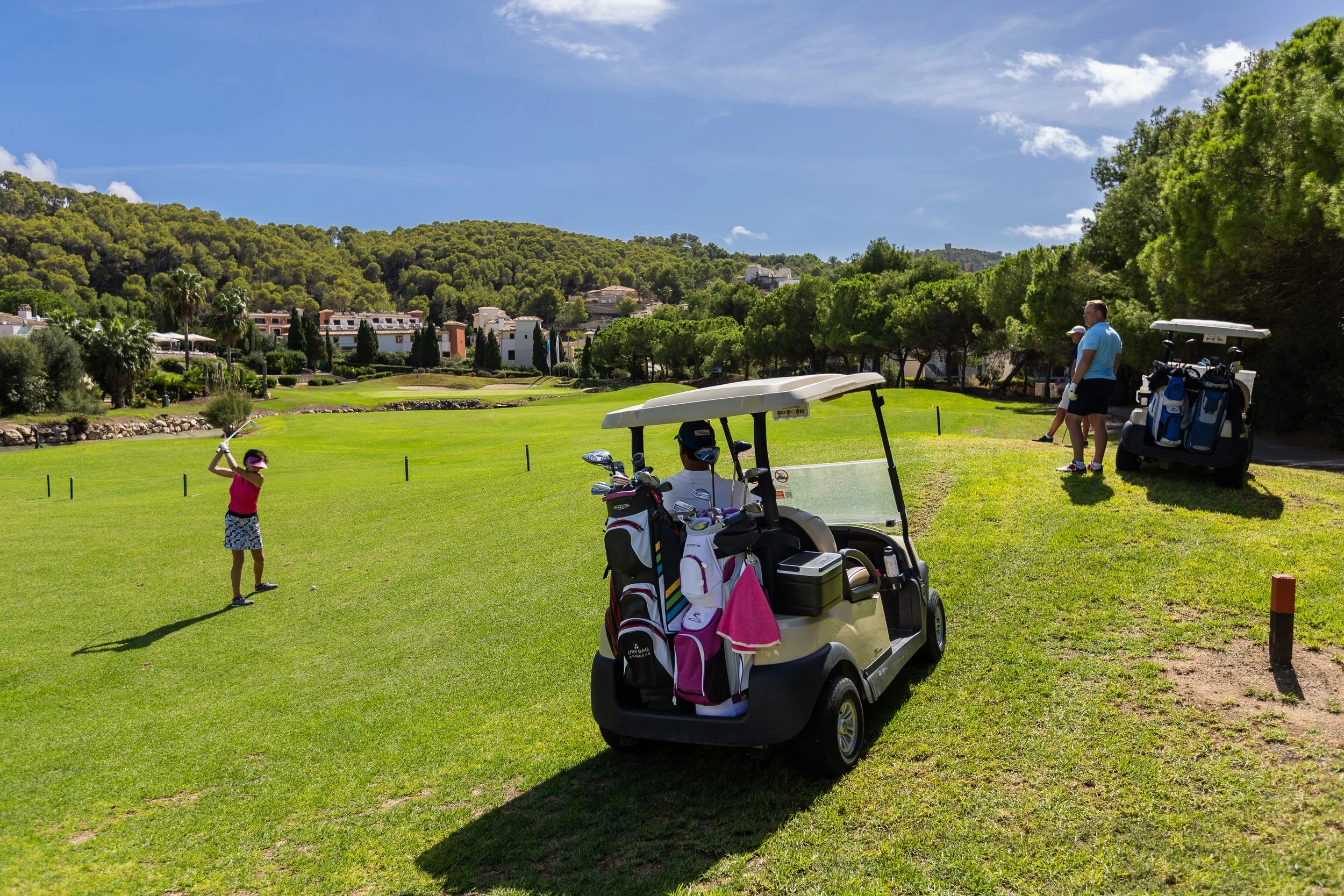 Niña jugando golf en un campo, dos adultos con carros de golf en la orilla y árboles en el fondo.