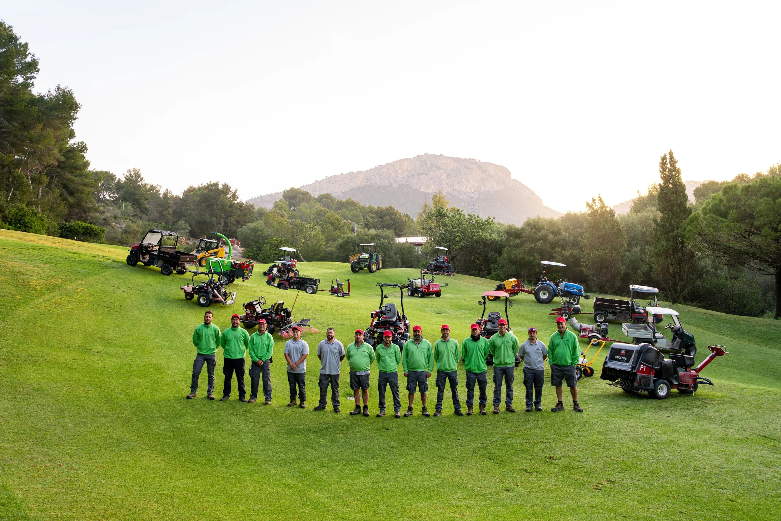 Grupo de diez personas en uniforme verde y gris, con gorra roja, de pie sobre césped en un campo de golf con varias máquinas cortacésped y equipos de mantenimiento en el fondo, rodeadas de árboles y montañas.