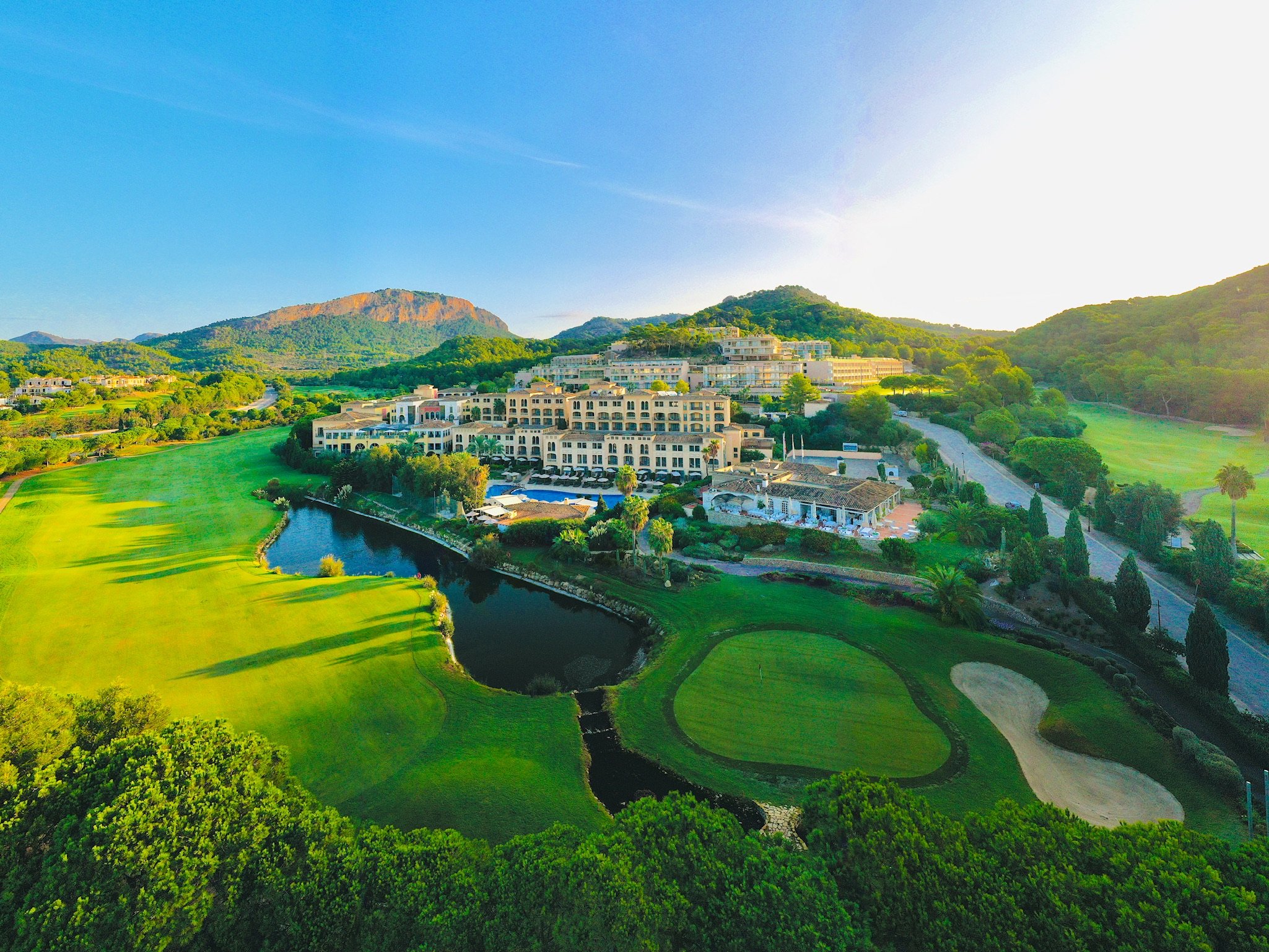 Vista aérea de un campo de golf con laguna, rodeado de árboles y edificios residenciales, con montañas al fondo bajo un cielo claro y soleado.