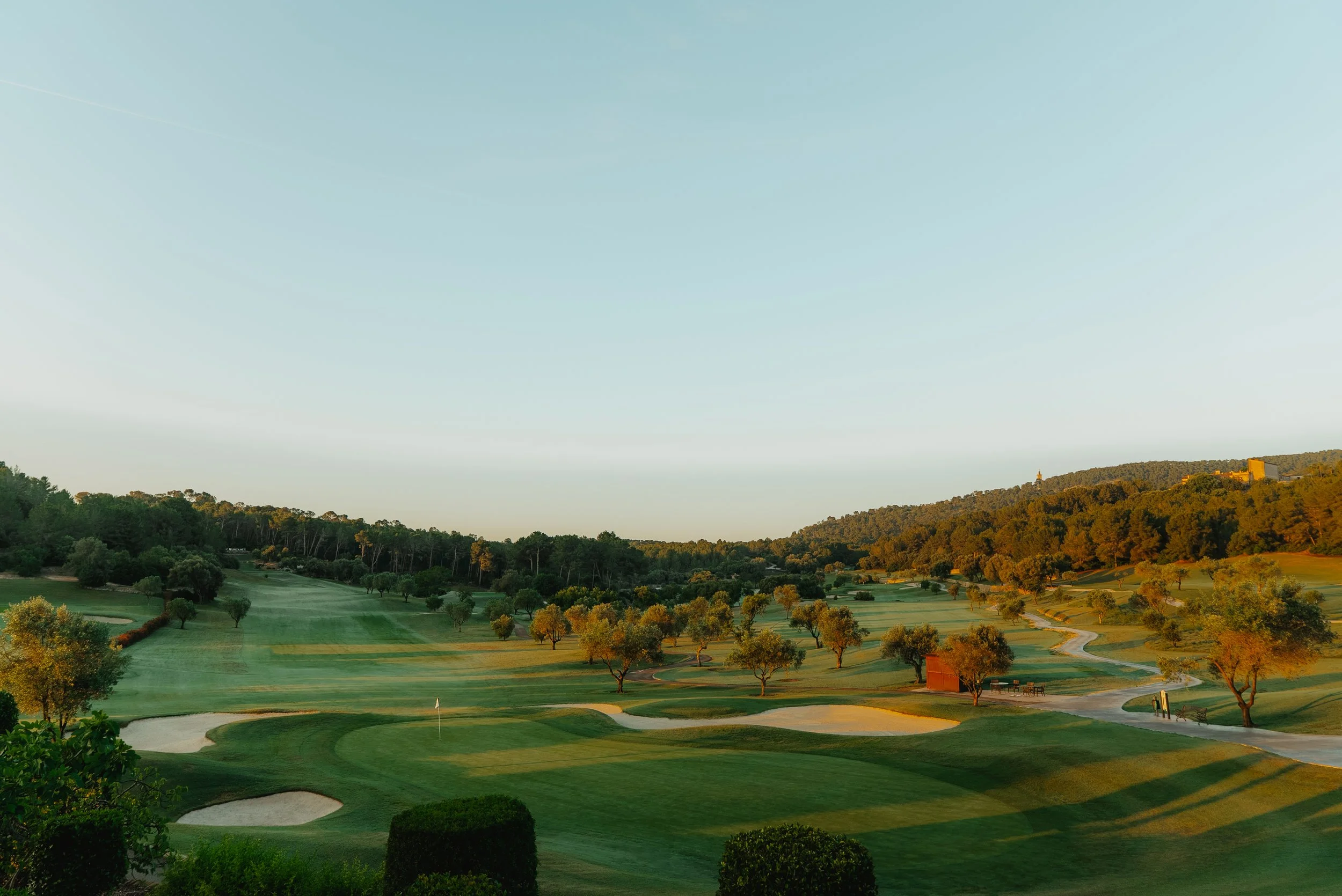 Vista panorámica de un campo de golf con césped bien cuidado, árboles dispersos, y colinas de fondo durante el atardecer.