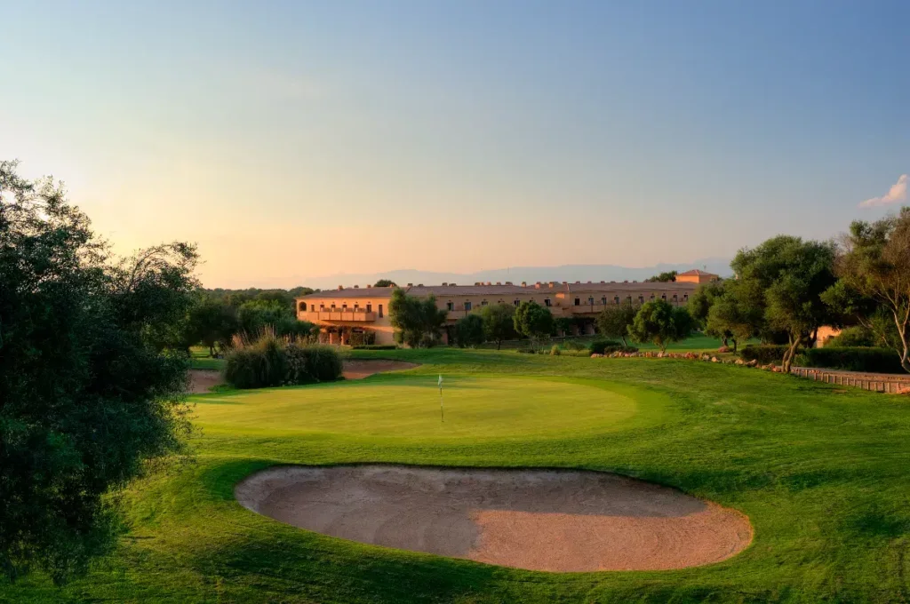 Vista de un campo de golf al atardecer con un green, un hoyo y un bunker, rodeado de árboles y un edificio en el fondo.