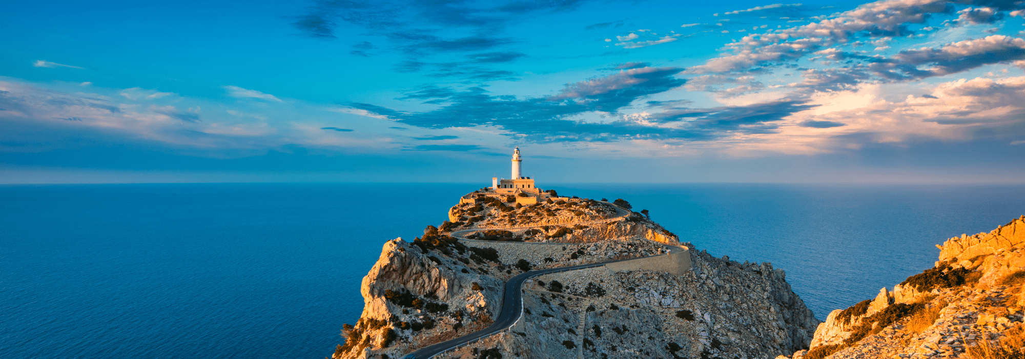 Faro en una isla, rodeado de mar, con camino que lleva hasta la estructura, durante atardecer o amanecer con cielo nublado.
