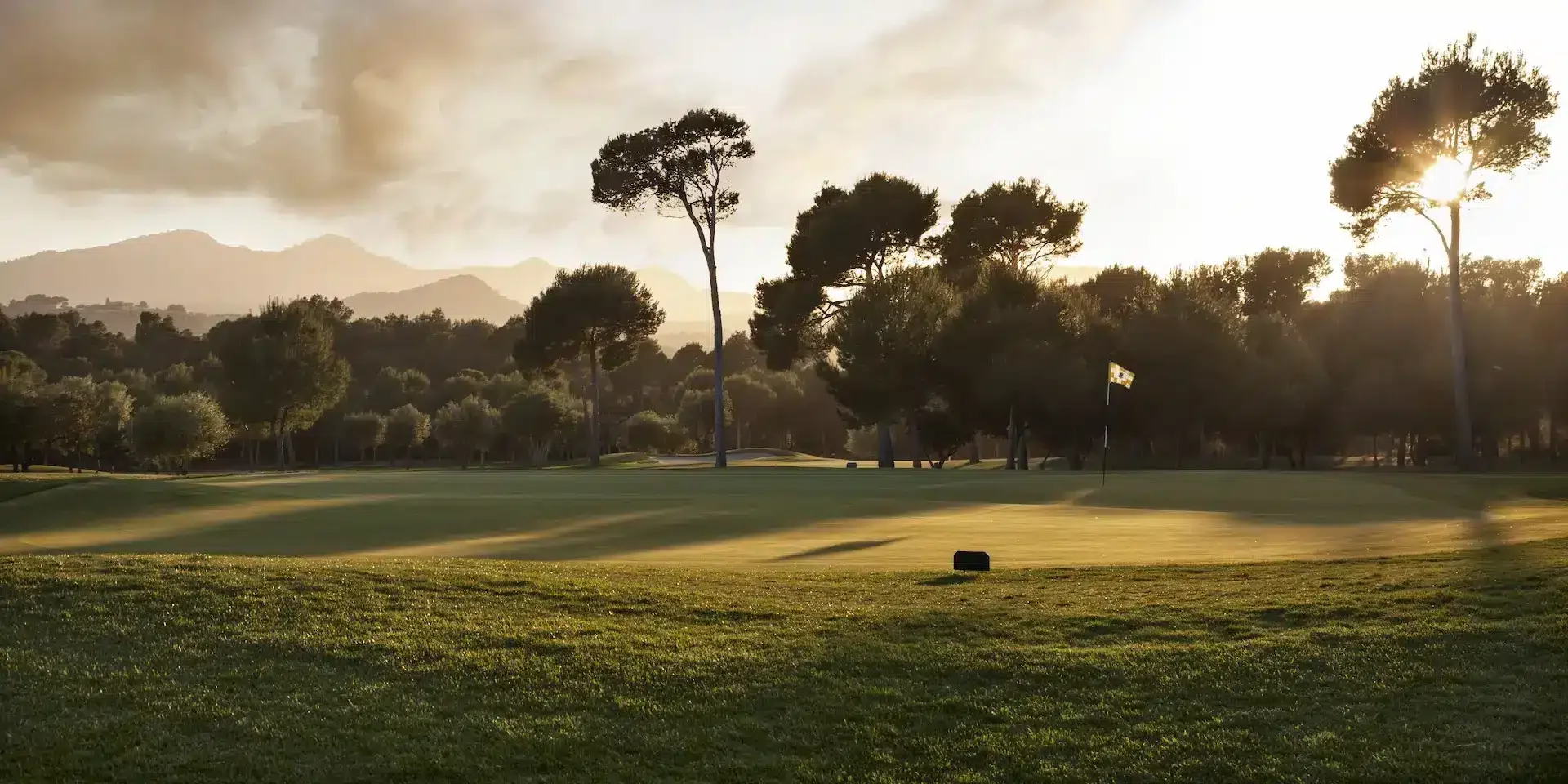 Campo de golf con bandera y árboles, al atardecer con montañas al fondo.