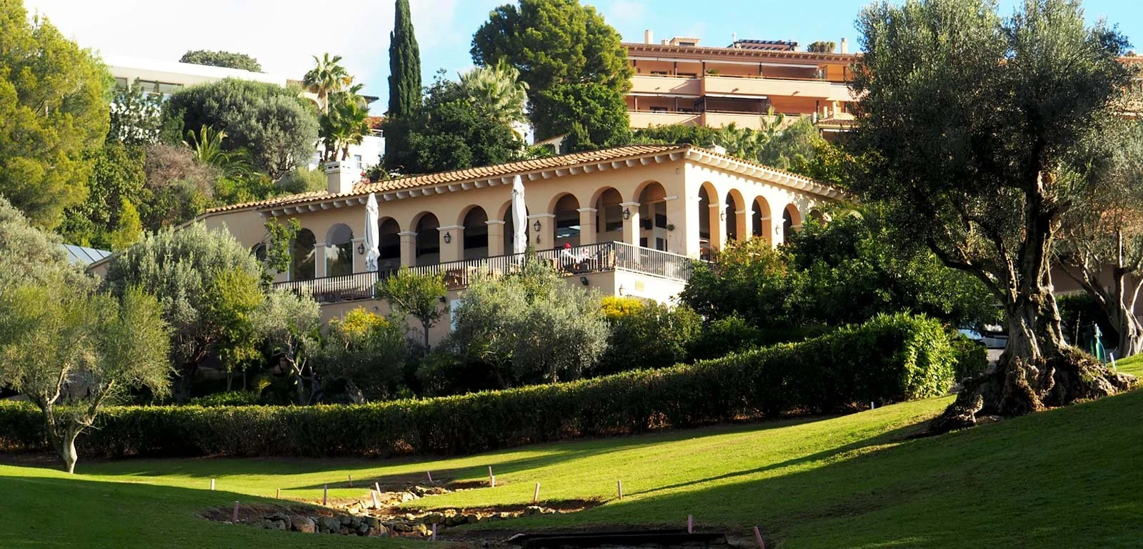 Casa de estilo mediterráneo con arco y terraza con sombrillas, rodeada de árboles y césped
