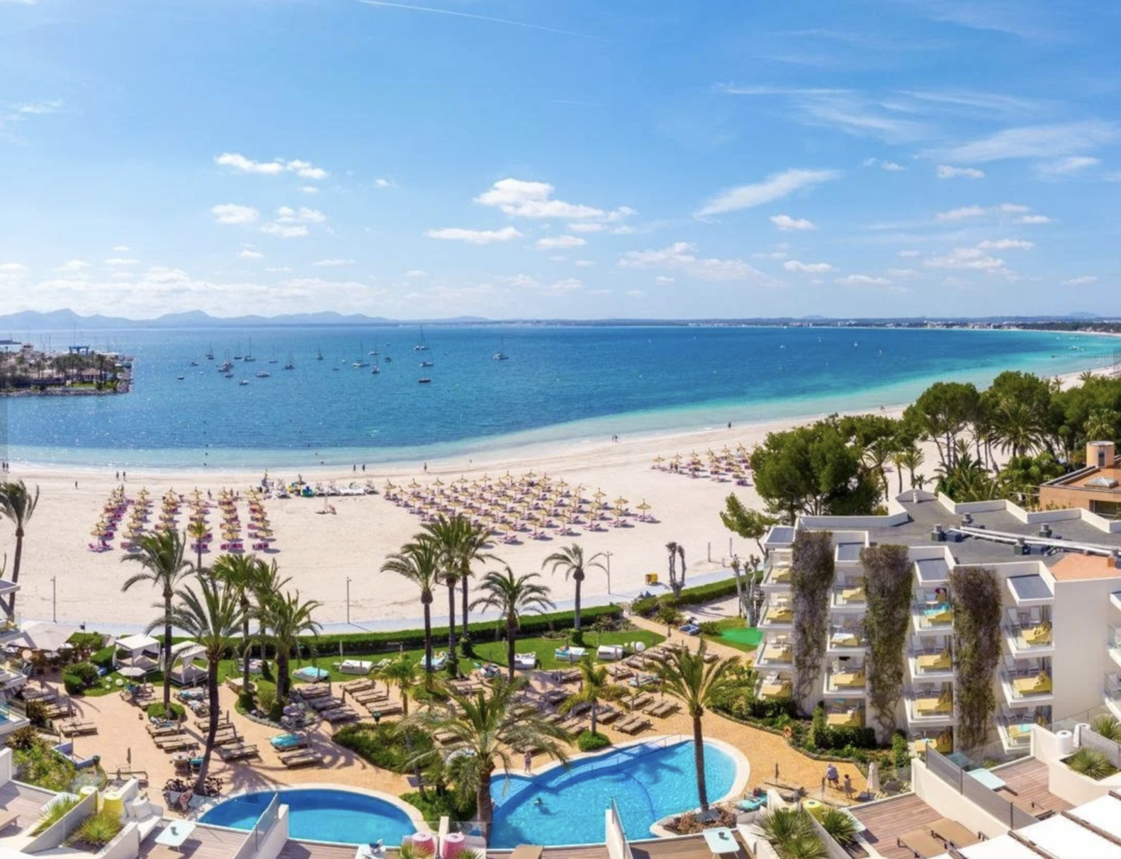 Vista panorámica de una playa con arena blanca, sombrillas y sillas, aguas azules y barcos en el mar, palmeras y un complejo hotelero con piscinas y edificios de colores claros.