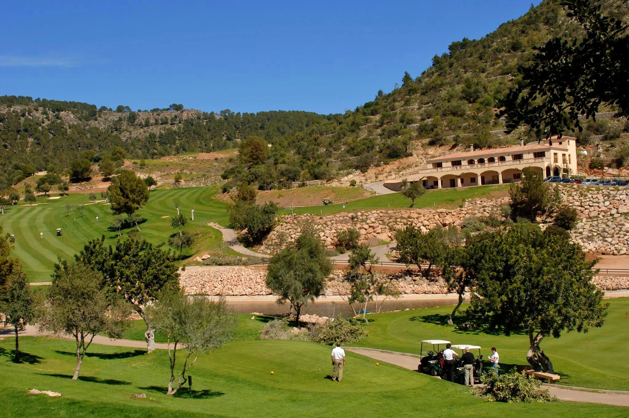 Vista de un campo de golf en un día soleado, con árboles y colinas, y en el fondo un edificio grande con terraza y una zona de estacionamiento.
