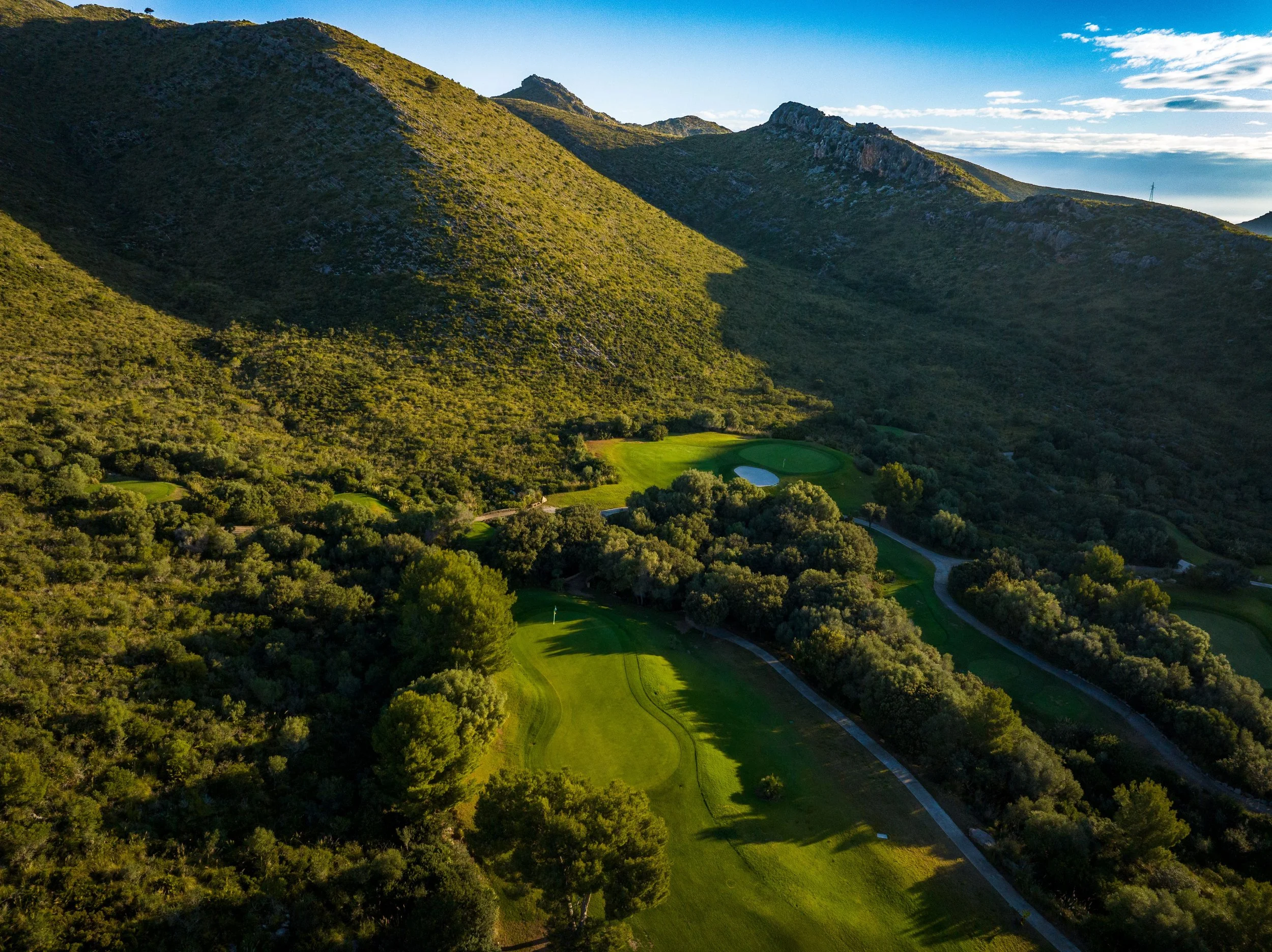 Vista aérea de un campo de golf en un valle rodeado de montañas verdes con cielo azul y algunas nubes.