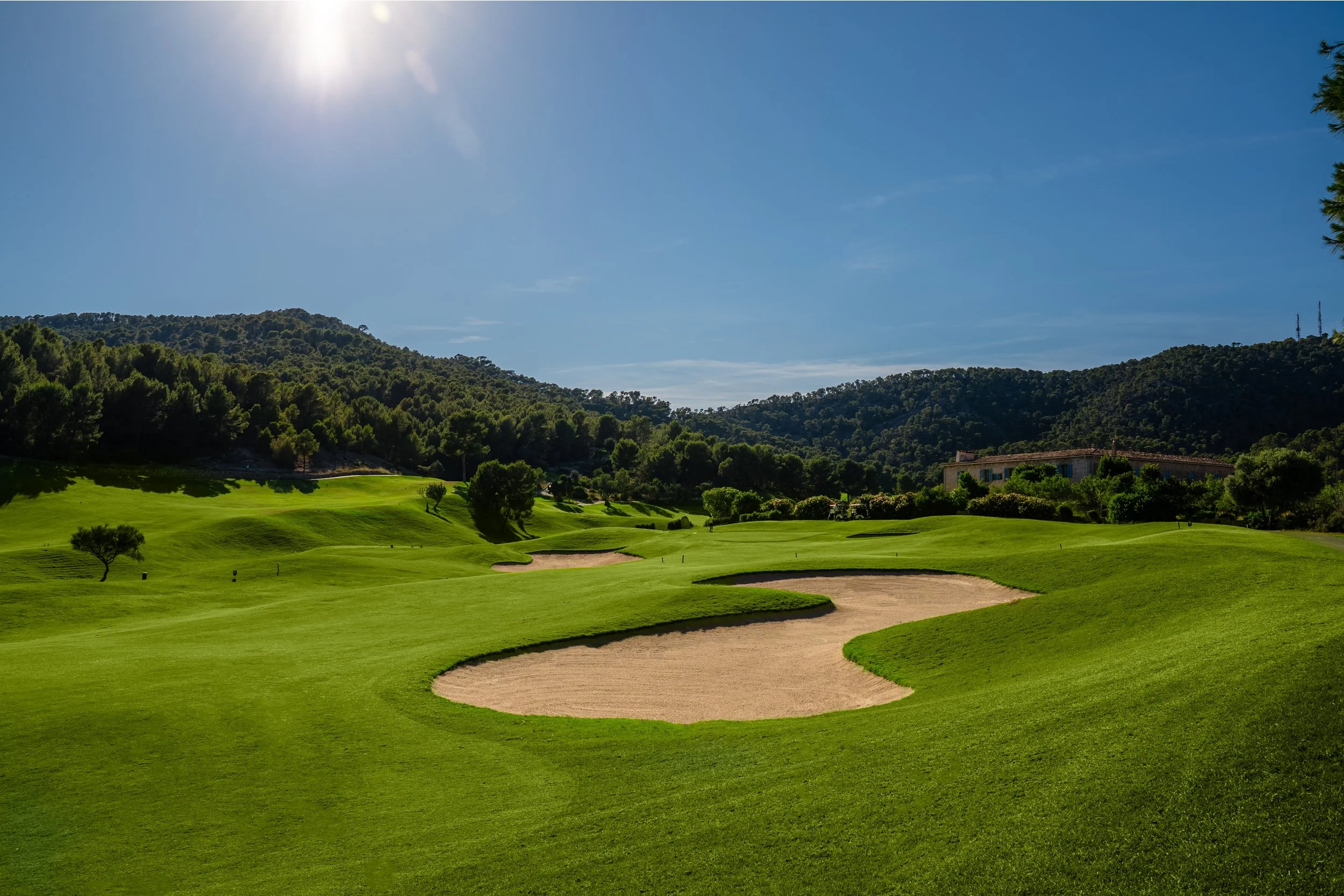 Campo de golf con pasto verde y dunas de arena, rodeado de árboles y colinas bajo un cielo azul con sol brillante.