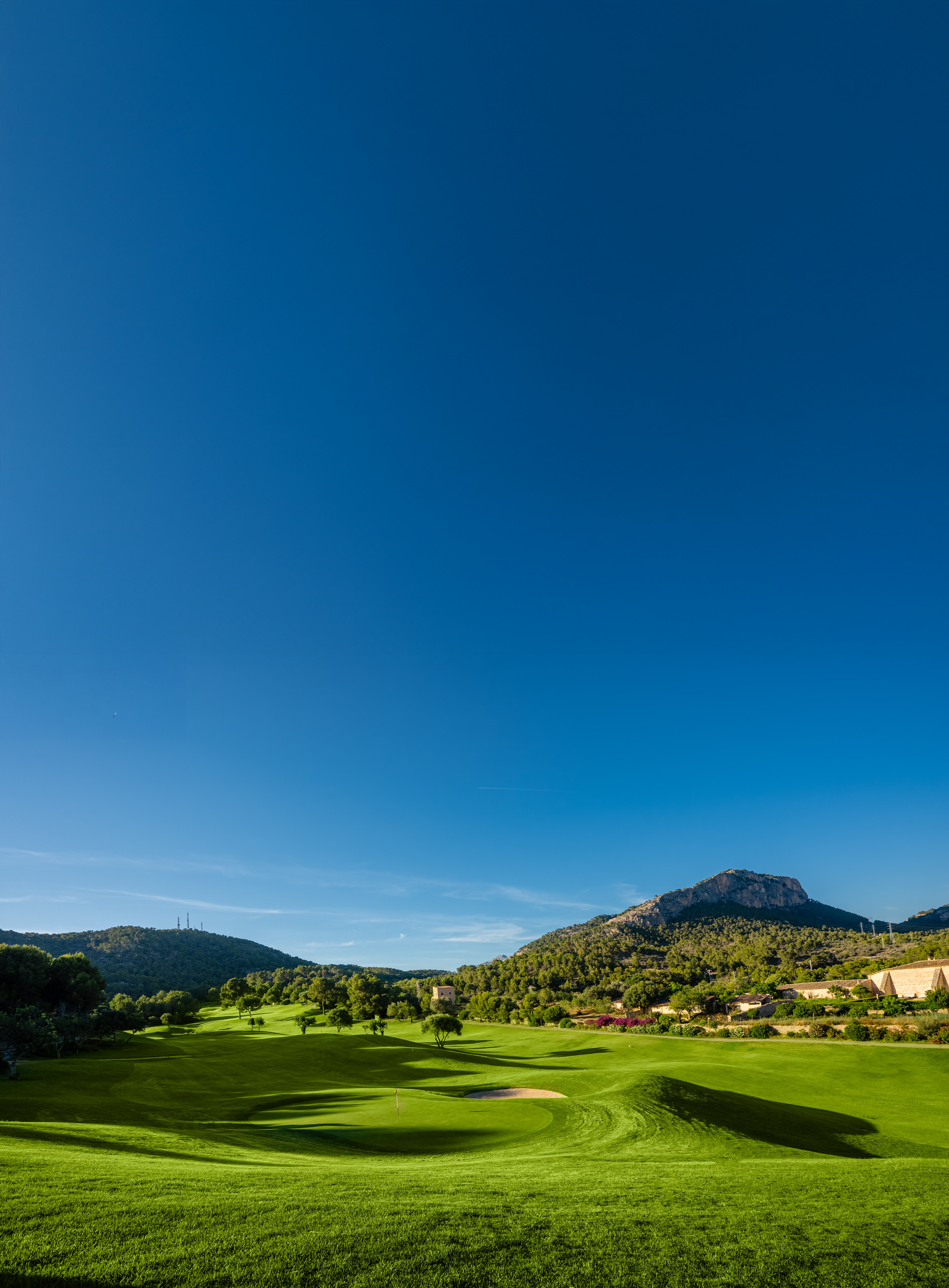 Campo de golf con césped verde, árboles dispersos, en un valle rodeado de montañas y un cielo despejado.