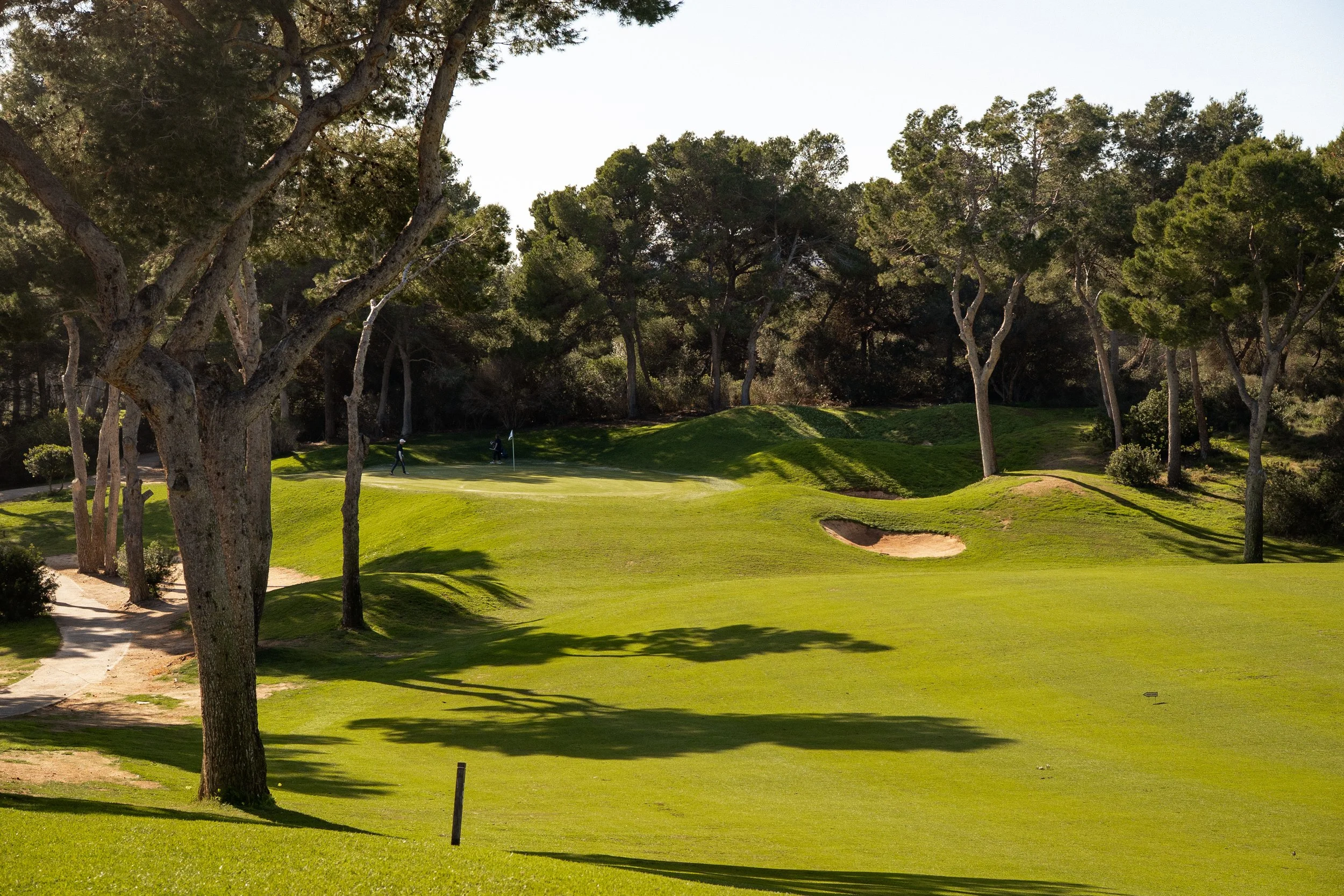 Paisaje de un campo de golf con árboles y césped verde, con un bunker y obstáculos de arena, y dos jugadores en el fondo.