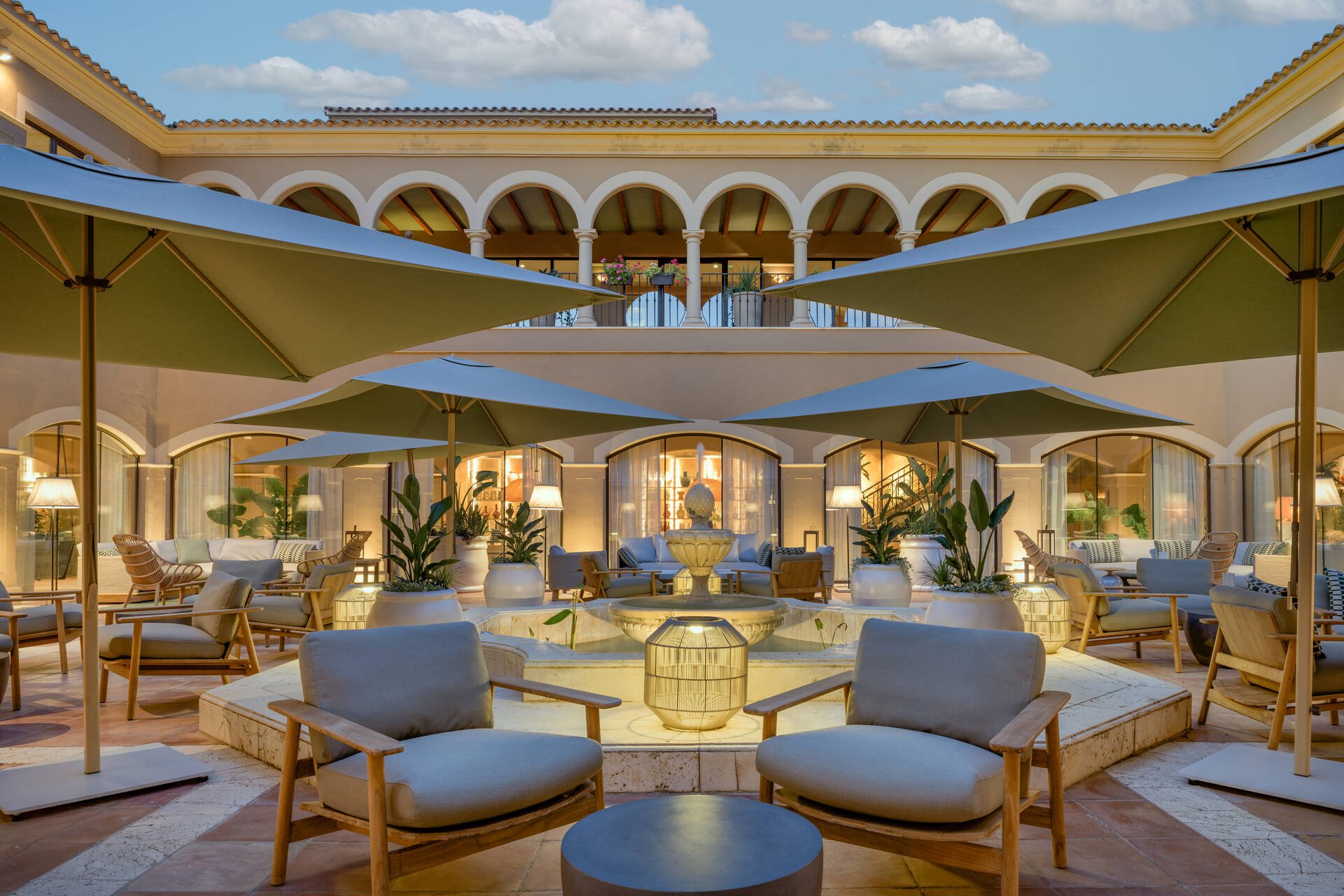 Un patio exterior elegante con sombrillas verdes, muebles de madera con cojines blancos, y una fuente en el centro. Al fondo, un edificio de varios pisos con arcos en el balcón y plantas en macetas.