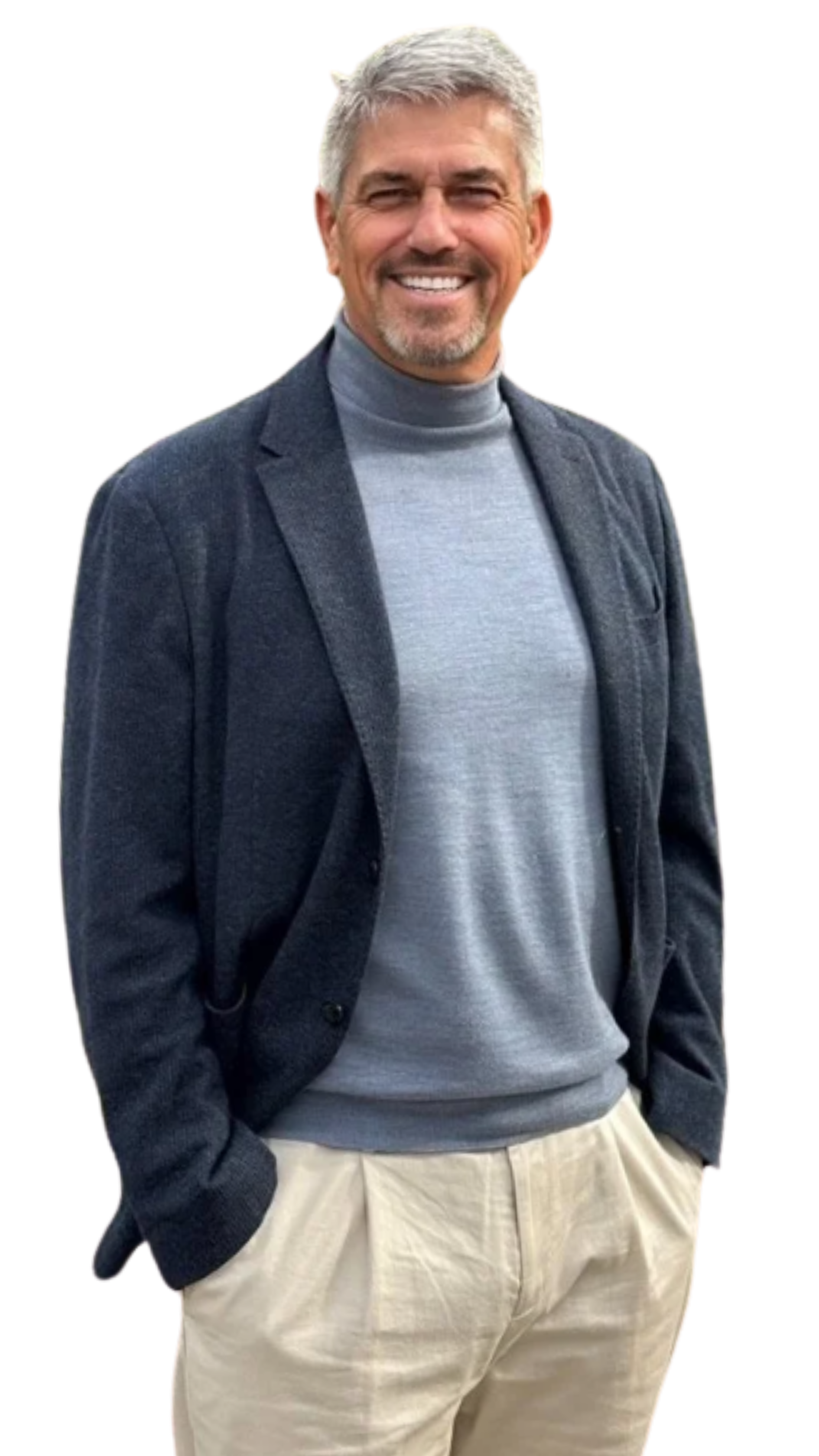 Hombre con cabello canoso y barba, sonriendo, viste suéter gris, chaqueta azul marino y pantalones claros.