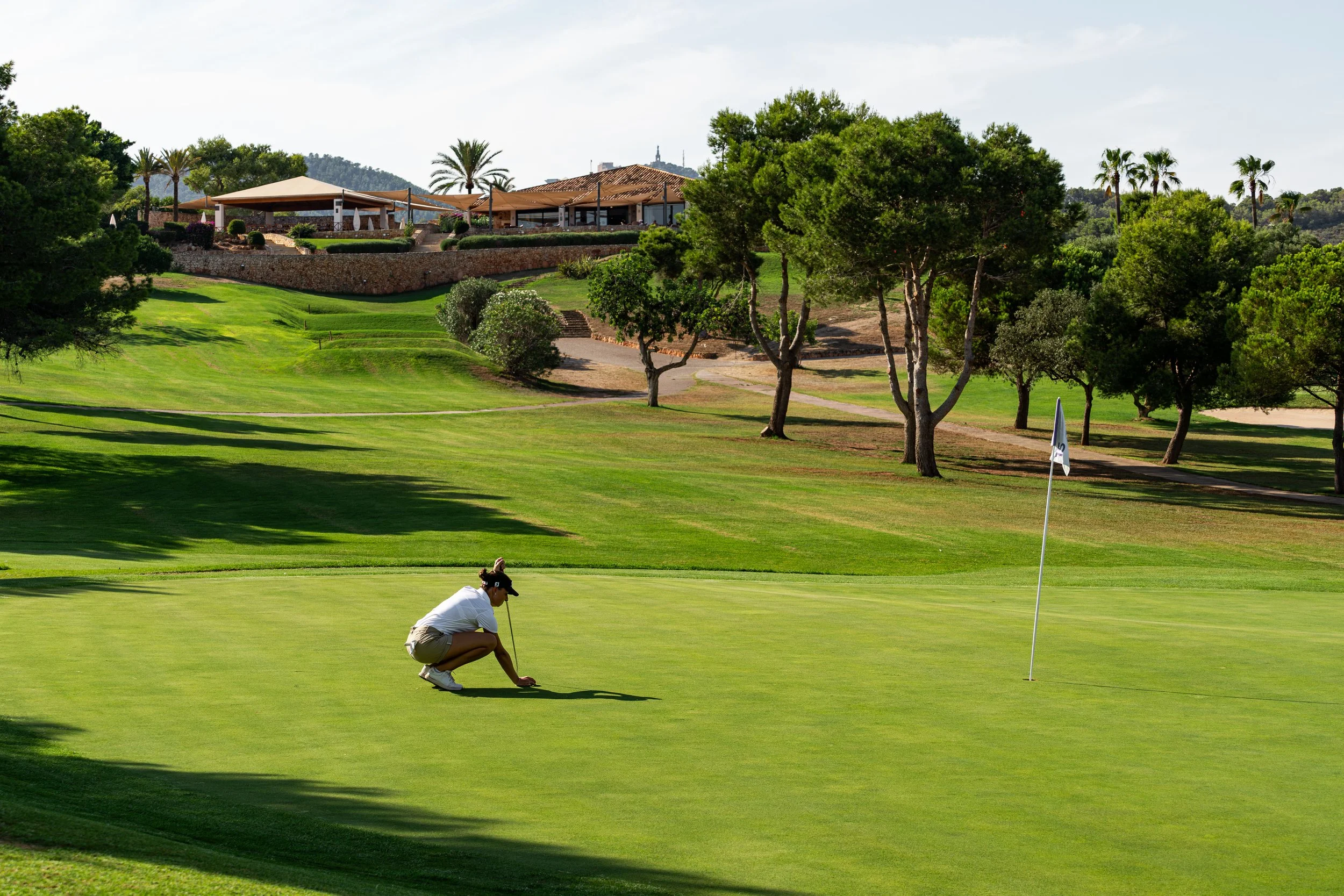 Persona jugando golf en el green en un campo con árboles y un edificio en la colina.