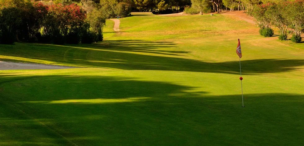 Cartel de golf en el hoyo de un campo de golf con césped bien cuidado, árboles y arbustos en el fondo, y sombras y luz del sol en el césped.