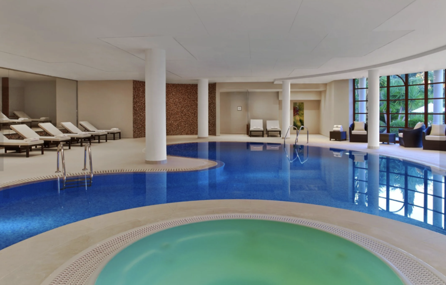 Piscina cubierta en un spa con tumbonas y áreas de descanso cerca de grandes ventanas con vista a plantas verdes.