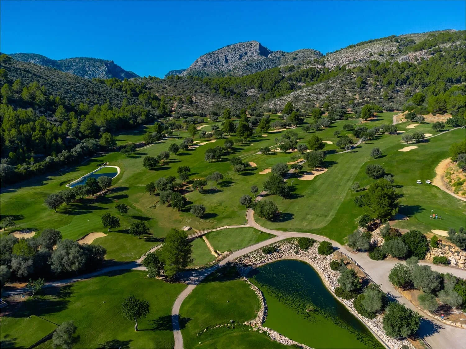 Campo de golf en un valle rodeado de colinas y montañas, con árboles dispersos, lagos y caminos peatonales