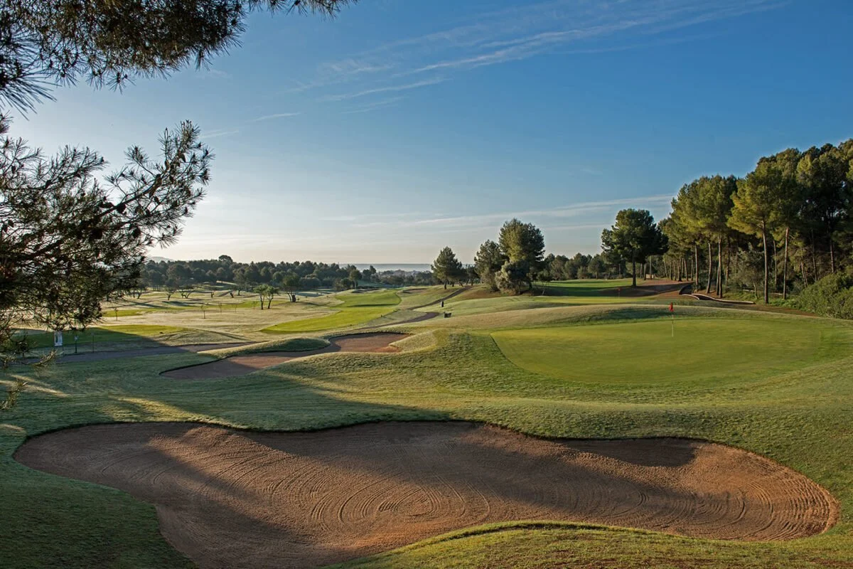 Campo de golf con hoyos y arena, rodeado de árboles y césped bajo un cielo despejado.