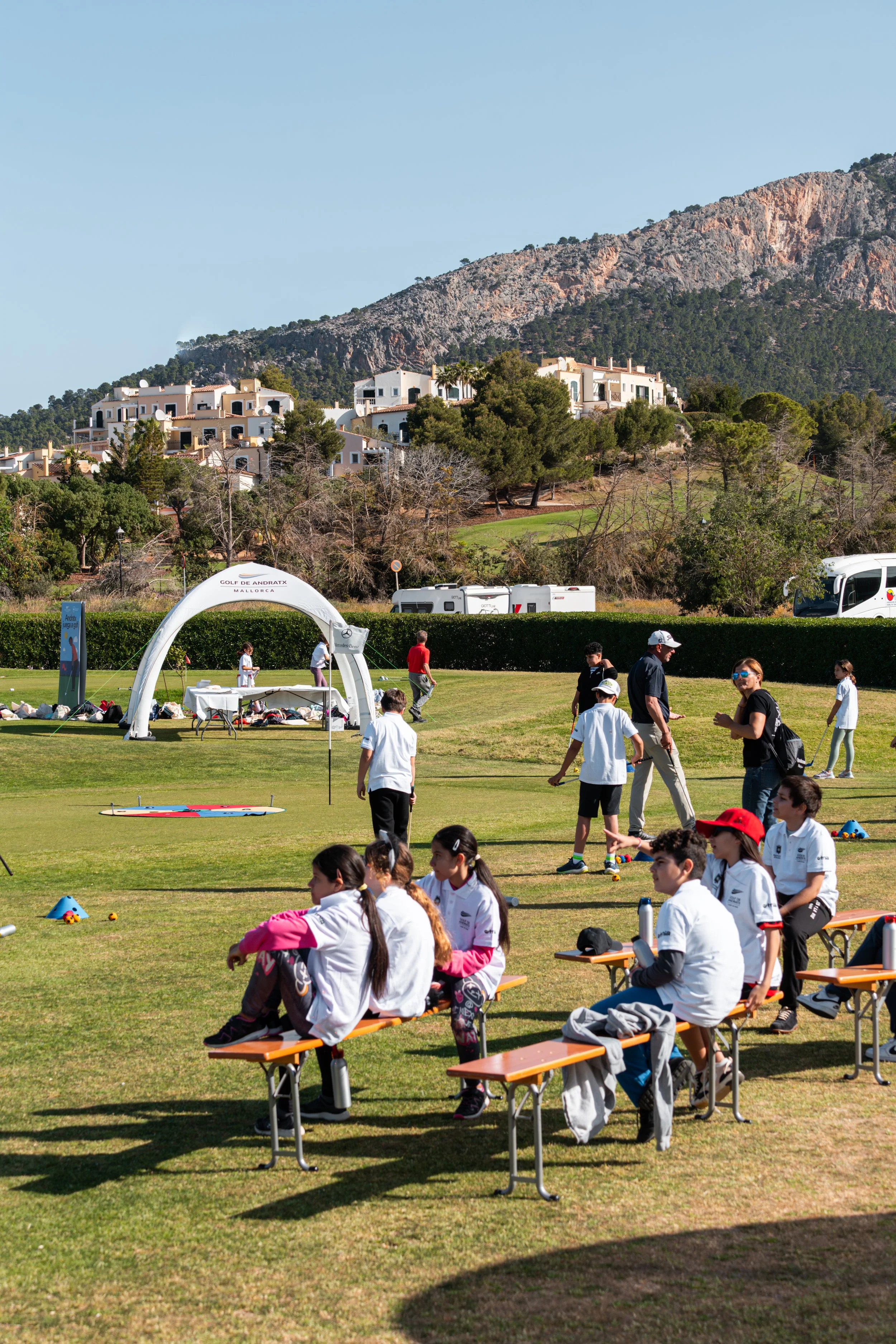 Niños y adultos en un campo de golf en un evento al aire libre en un día soleado con montañas al fondo.