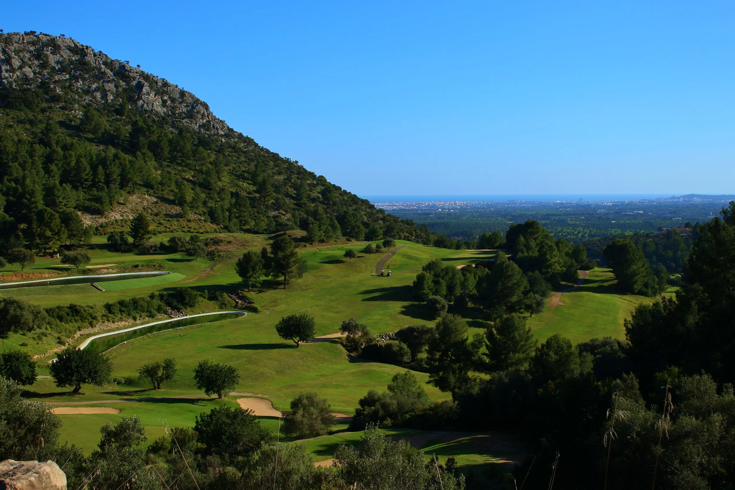 Vista panorámica de un campo de golf rodeado de árboles, con una colina en el lado izquierdo y el océano en el horizonte bajo un cielo despejado.
