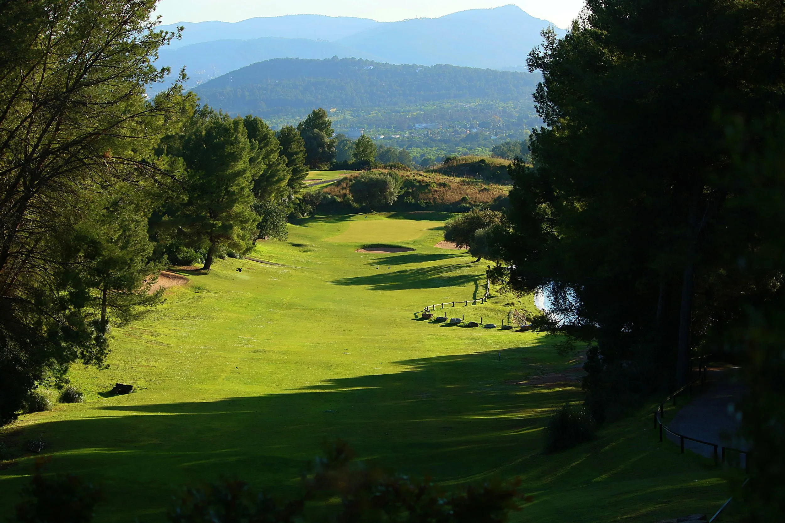 Un campo de golf rodeado de árboles verdes, con montañas al fondo y un lago pequeño a la derecha.
