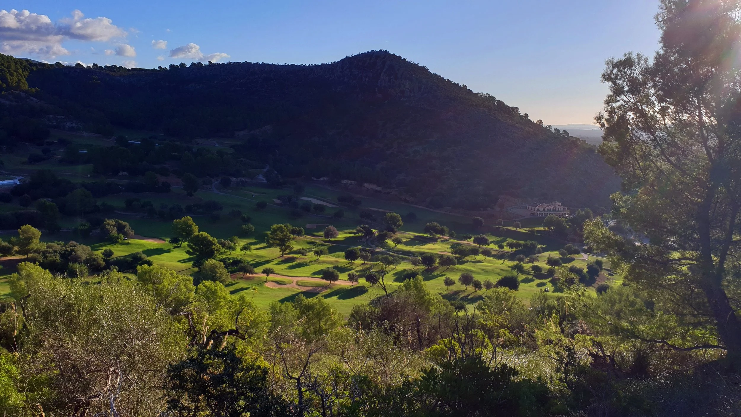 Vista panorámica de un campo de golf rodeado de árboles y colinas, con una montaña al fondo y el sol brillante en el cielo.