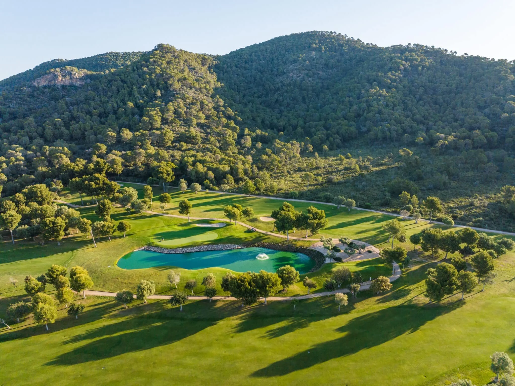Vista aérea de un campo de golf con un lago, rodeado de árboles y colinas verdes.