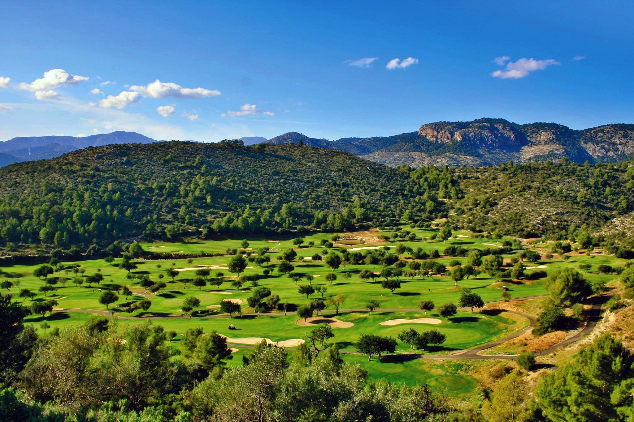 Vista panorámica de un campo de golf rodeado de colinas y montañas con cielo despejado y algunas nubes.