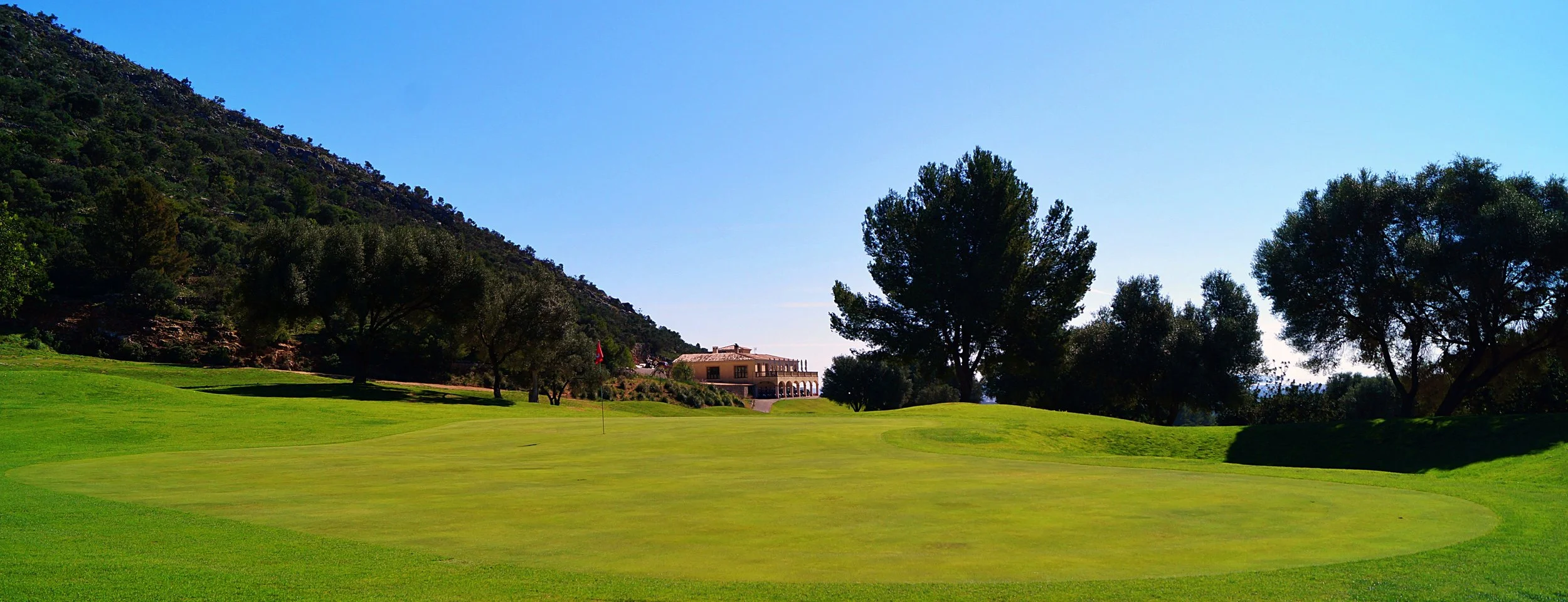 Campo de golf con green, bandera, árboles y una casa en el fondo, rodeado de montañas y cielo despejado.