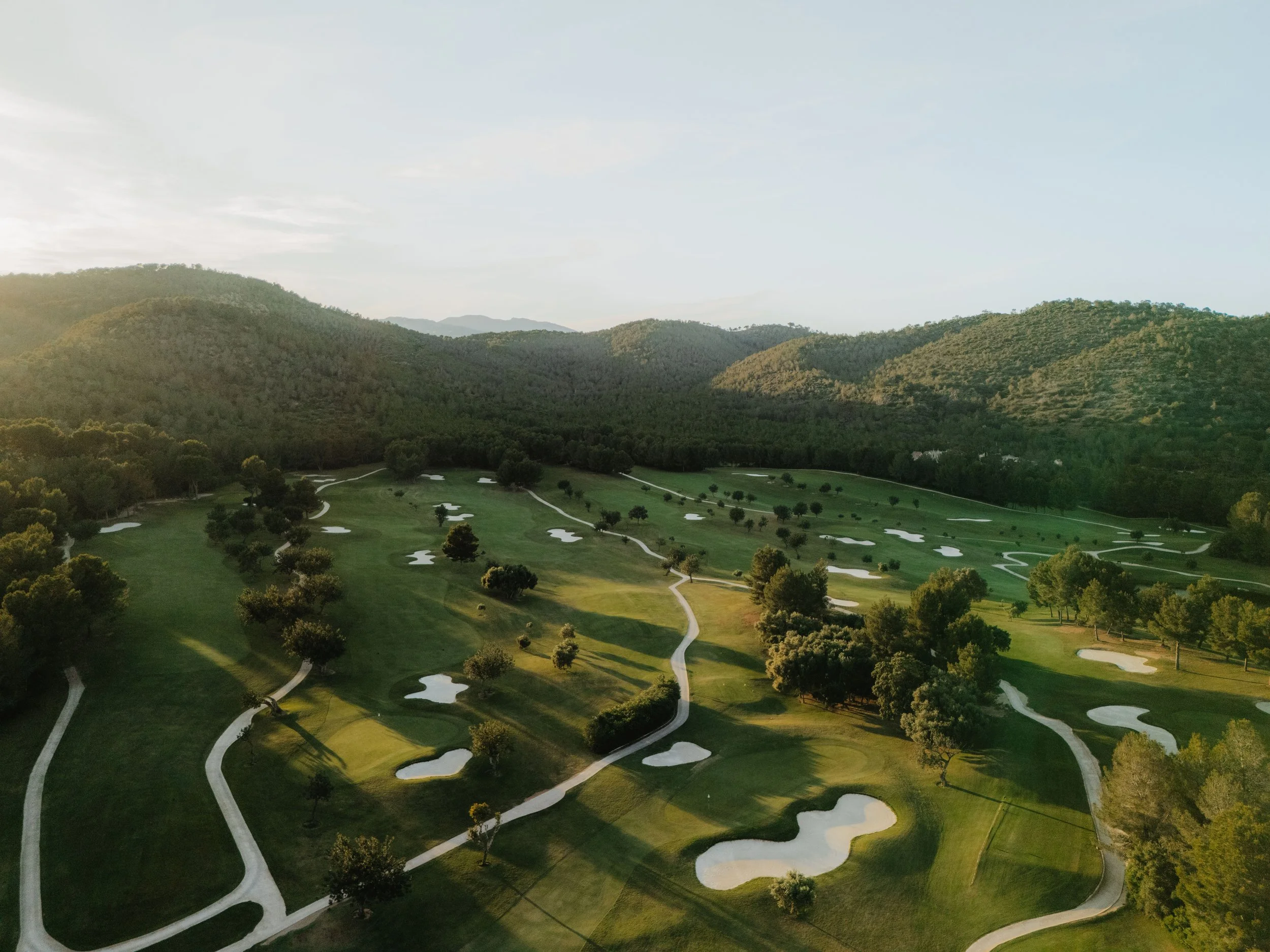 Vista aérea de un campo de golf con caminos, árboles y bunkers, rodeado de montañas y con cielo despejado.