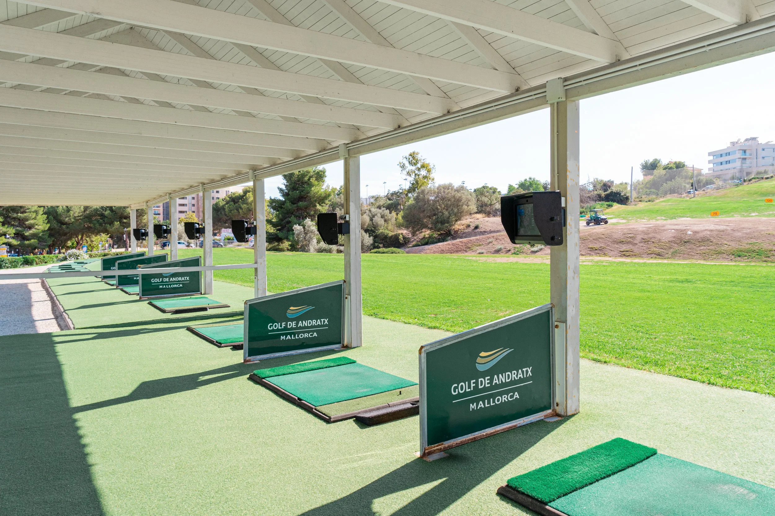Zona de práctica de golf en Mallorca, con varias estaciones de tiro y pantallas de monitoreo bajo un techo cubierto, rodeada de áreas verdes y árboles.