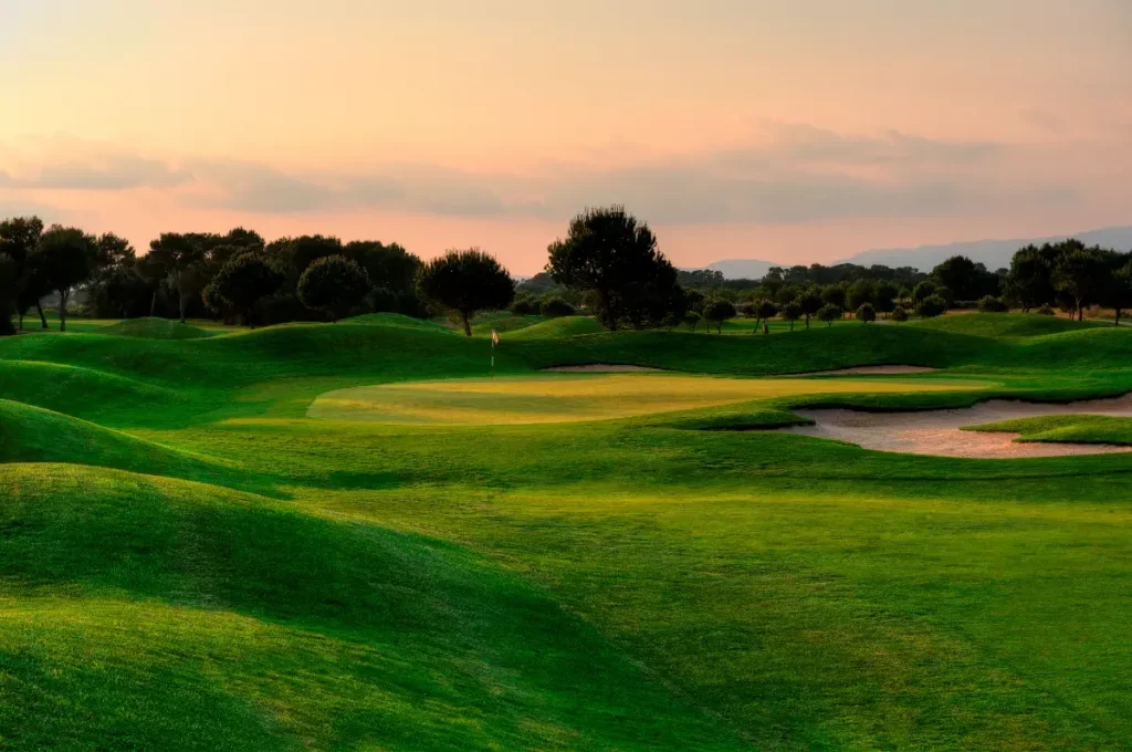 Campo de golf con césped verde, árboles dispersos y búnkeres, al atardecer con cielo nublado en el fondo.