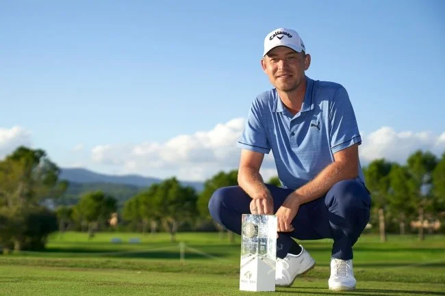 Jugador de golf en un campo de golf, sosteniendo un trofeo, usando gorra y ropa deportiva, con cielo despejado al fondo.